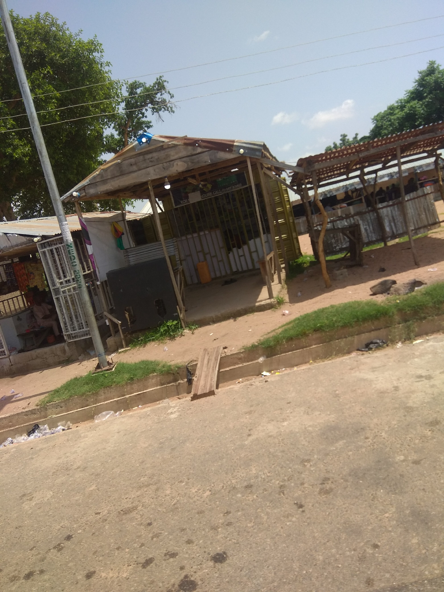 The image depicts a street scene in Jalingo, Nigeria, characterized by a roadside market stall and its surroundings. The stall, constructed with a corrugated metal roof and wooden supports, displays various items for sale, including what appears to be clothing. A person is seated near the stall, possibly a vendor or customer. Adjacent to the stall, a large, dark, rectangular object, possibly a speaker or a generator, is visible. The ground is a mix of dirt and paved road, with some scattered debris and vegetation. The environment suggests a typical urban setting in Nigeria, with the presence of market activities and common roadside features.