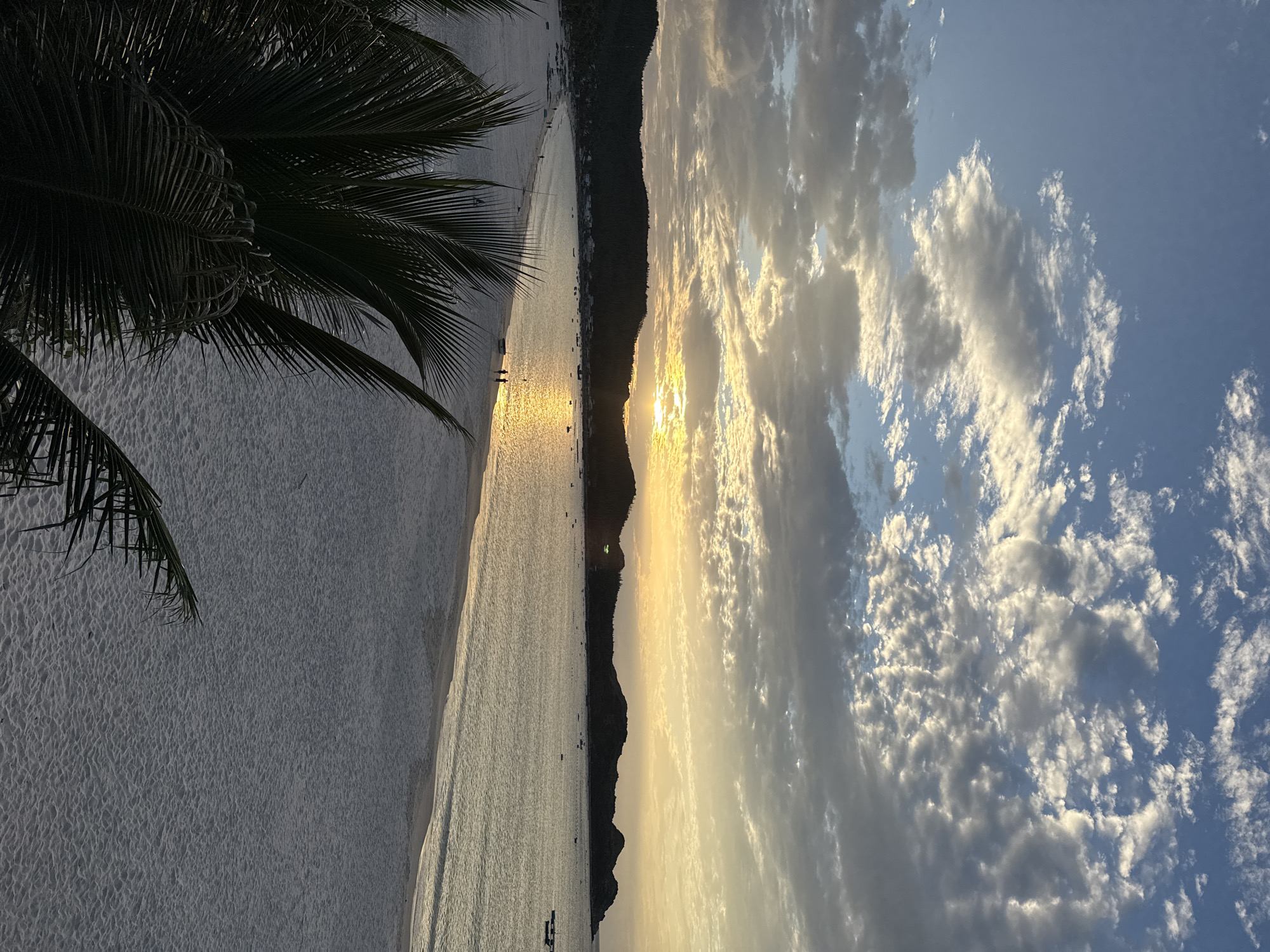 The image depicts a serene sunset over a tranquil beach in Song Cau, Vietnam. The sun, a brilliant orb of golden light, is low on the horizon, casting a warm glow across the sky and reflecting on the calm sea. The sky is adorned with a scattering of fluffy white and gray clouds, tinged with orange and yellow from the setting sun.
In the foreground, the dark silhouettes of palm fronds frame the view, adding a tropical touch to the scene. The beach itself is a vast expanse of soft, white sand, stretching along the coastline. Along the shore, a few scattered individuals can be seen walking and interacting, their figures small against the immensity of the landscape.
Beyond the beach, a chain of dark, undulating hills forms the backdrop, dotted with a few buildings and small boats anchored in the bay. The overall atmosphere is one of peace and natural beauty, capturing the essence of a coastal sunset in Vietnam.