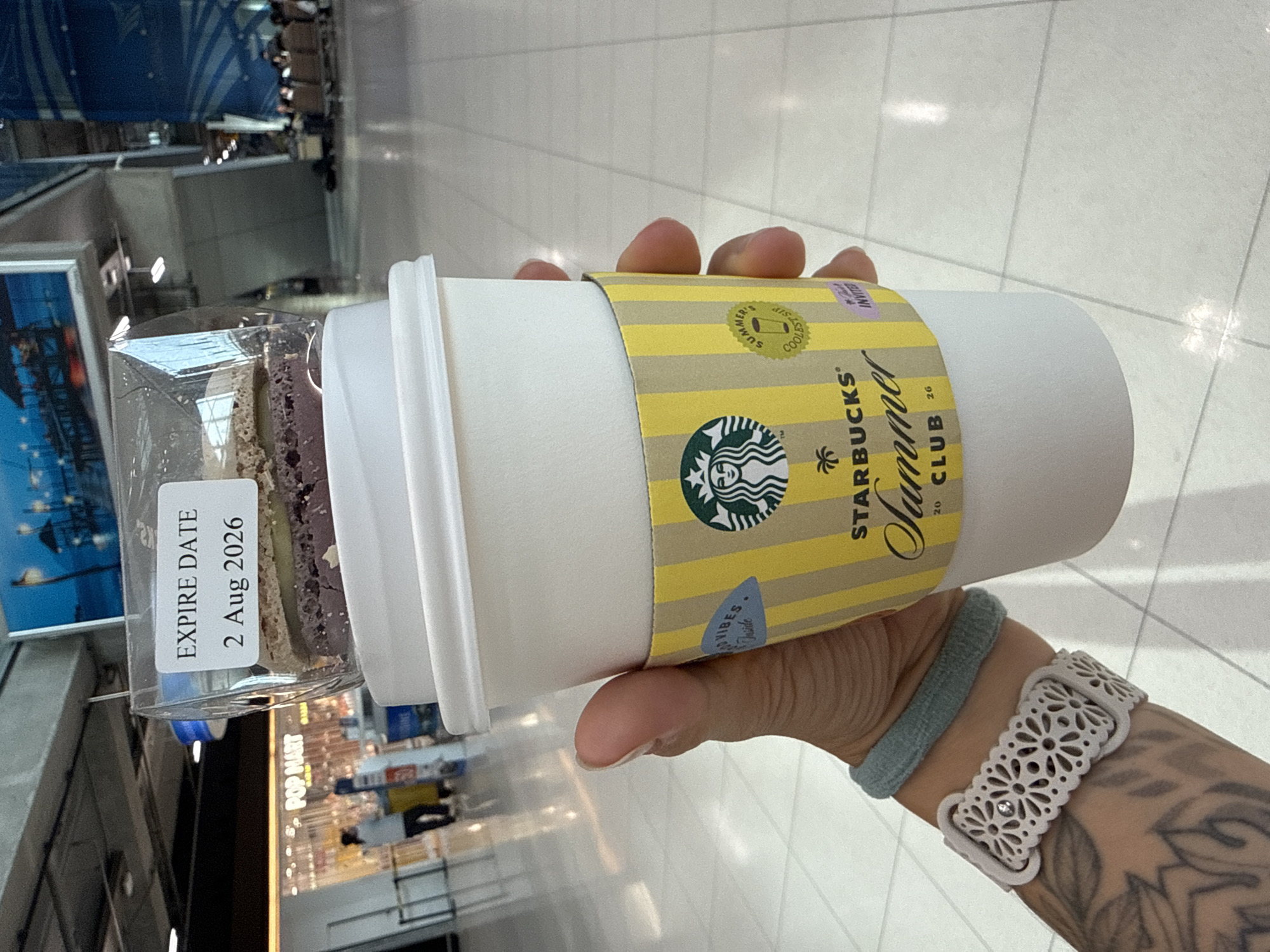 A person's hand is holding a Starbucks coffee cup with a "Summer Club" sleeve at what appears to be an airport. A clear plastic container with a dessert is resting on top of the cup, displaying an "EXPIRE DATE 2 Aug 2026" label. In the background, a "POP MART" storefront is visible, and people are seen in the distance in a well-lit, tiled area. The overall environment suggests an indoor public space like an airport terminal.
