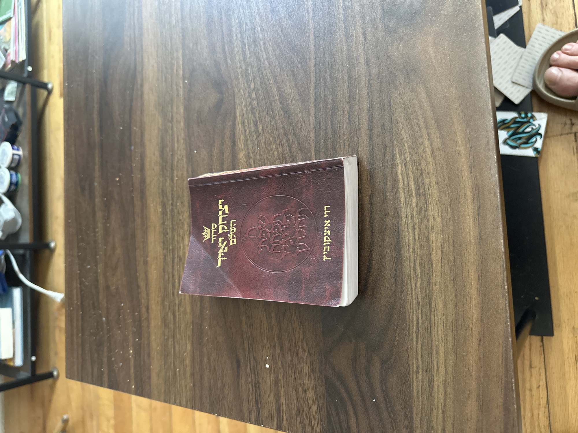 A medium shot depicts a brown prayer book lying on a brown wooden table. The book is oriented so that its spine and cover are visible. The cover is a reddish-brown color and features a circular design in the center. Hebrew lettering is visible on the cover. The table has a smooth, polished surface with wood grain visible. In the upper right corner of the image, a glimpse of a sandal-clad foot and various items on the floor can be seen. A metal frame with assorted objects on shelves is visible on the left side of the frame. The overall lighting appears to be ambient and natural, giving the scene a warm tone. There is no explicit indication of the location being in New York City, United States of America.