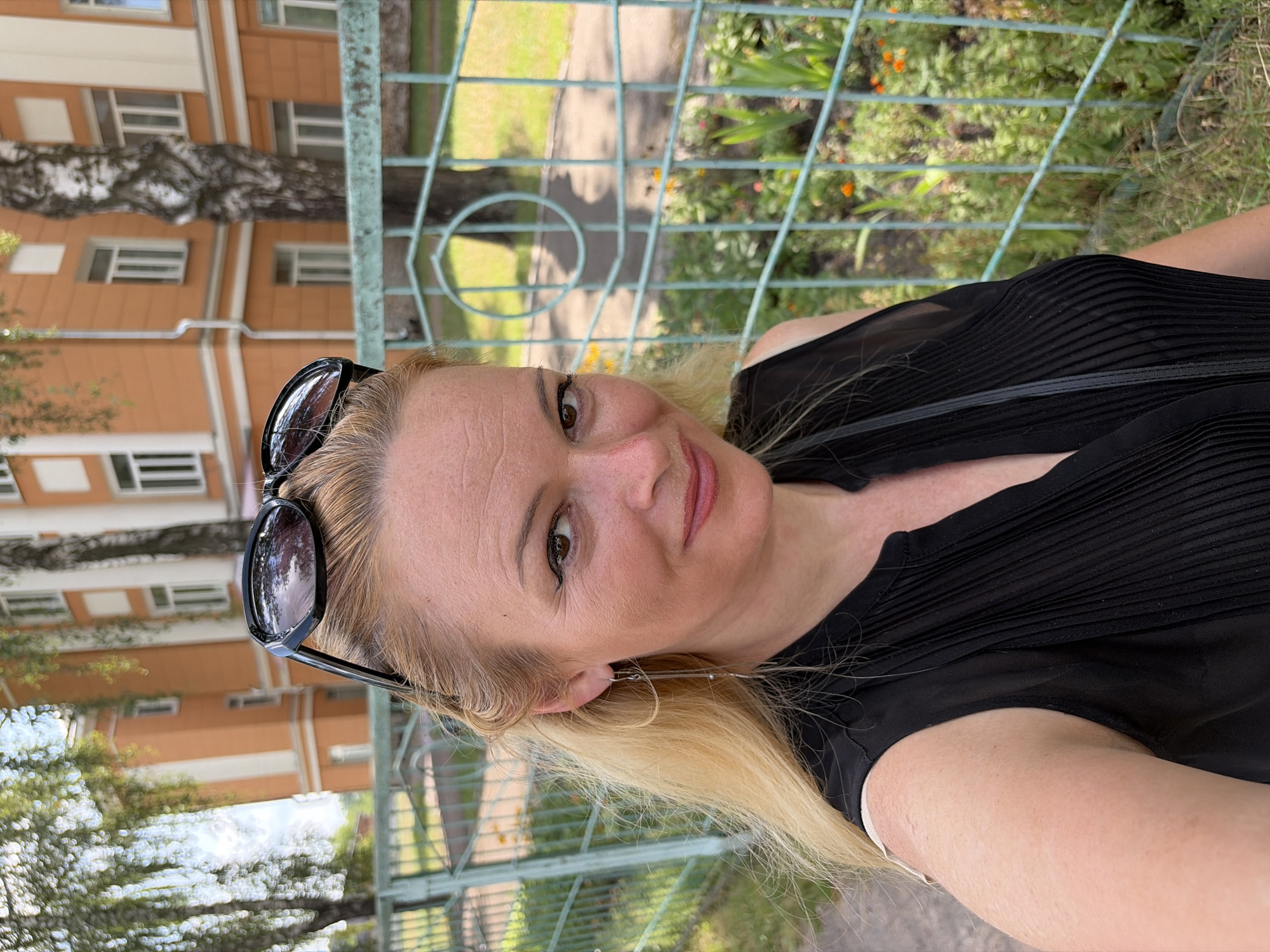 A blonde woman with sunglasses perched on her head stands in front of a light orange building with white trim. She is wearing a black sleeveless shirt. Behind her, a green metal fence encloses a patch of grass with small orange flowers and green plants. Trees with light-colored bark are visible behind the building. The lighting suggests it is daytime, with shadows indicating the sun is present.