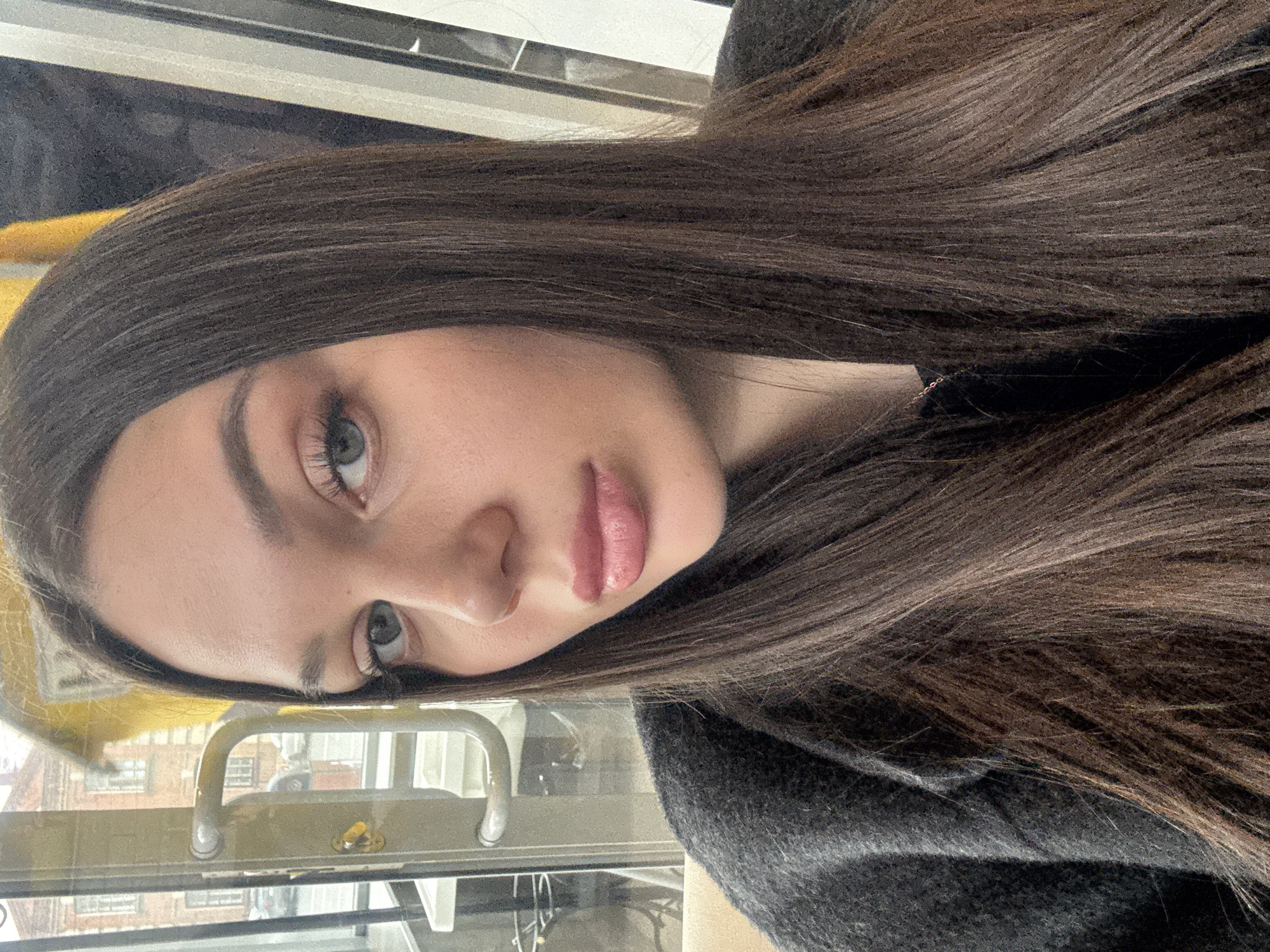 A young woman with long, straight brown hair and light eyes is looking directly at the camera. Her lips are pink and slightly parted. She is wearing a dark gray sweater and appears to be in some kind of public transport vehicle, possibly a bus or train, as indicated by the metal handrail and the window with an urban scene visible through it. The city is of Unknown, Unknown.