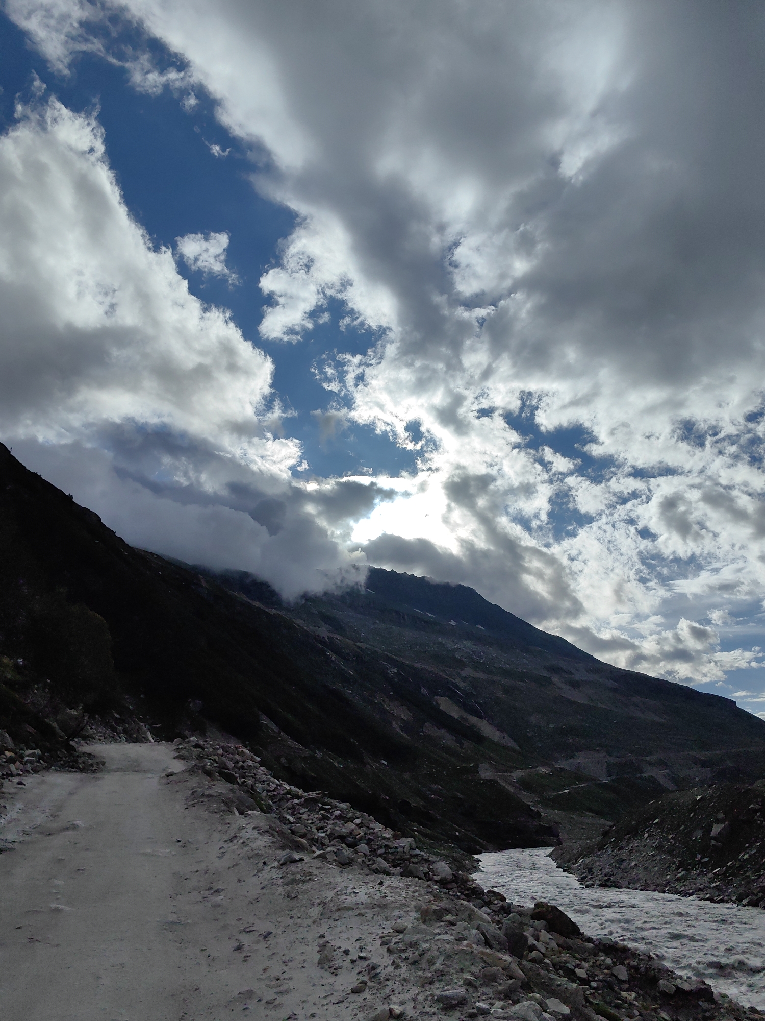 This image depicts a rugged, mountainous landscape with a dirt road winding along the left side. A fast-flowing river runs through the valley on the right, bordered by rocky terrain. The sky is filled with dramatic, swirling clouds, with patches of blue visible and sunlight breaking through. The overall scene suggests a remote, natural environment. The image does not provide information to locate it in Burwa, India.