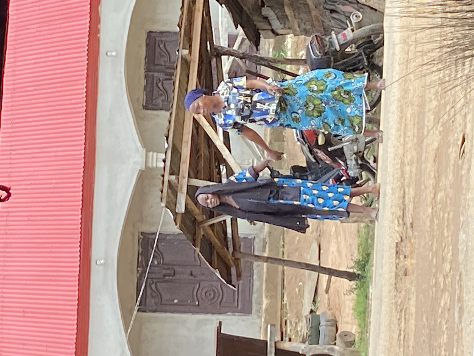 Two women stand near a building with a corrugated pink roof and arched entrances in Mararaba, Nigeria. The first woman wears a blue head covering and a dress with blue and green floral patterns, standing beside a motorcycle. The second woman wears a blue patterned dress with a black covering. The building has wooden supports and a dark door with ornamental features. A dirt road is visible, with some greenery and an object resembling a grinder near the edge of the road.
