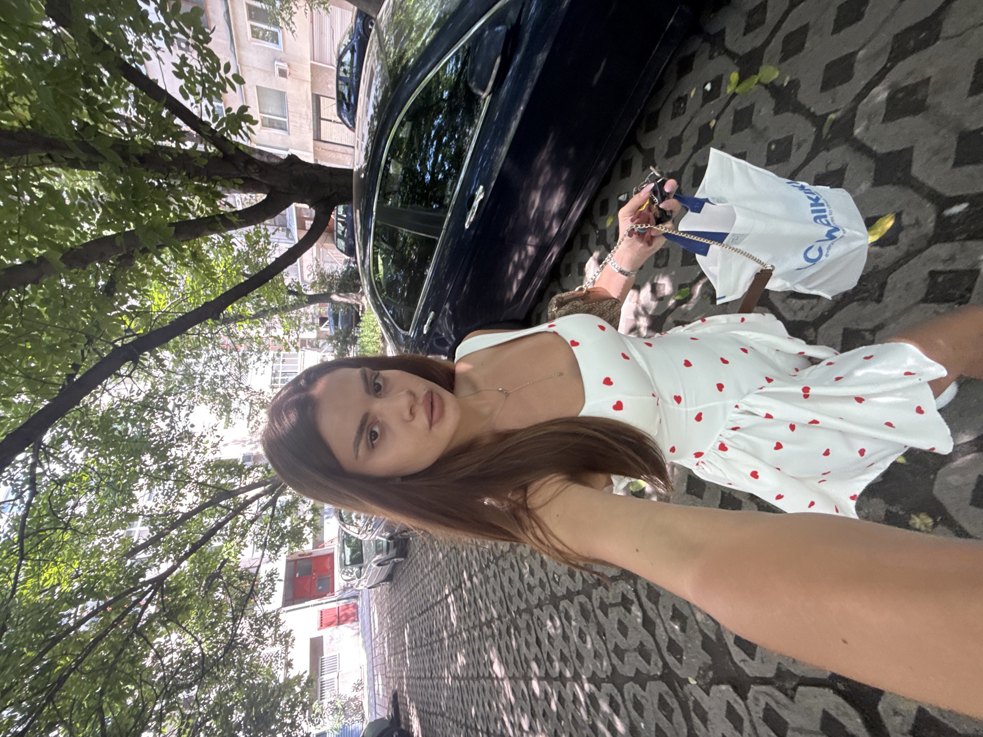 A young woman is depicted in a selfie-style outdoor photograph within an urban environment. She has long, straight, dark brown hair and is wearing a white, sleeveless dress with numerous small red heart patterns. A thin, gold-colored necklace and a silver-colored bracelet are visible. She holds a small, light brown patterned handbag with a chain strap over her left wrist and, in her left hand, a set of keys with a black fob and a white plastic shopping bag featuring a blue "LC Waikiki" logo and text.
The ground is paved with interlocking concrete blocks in varying shades of gray, displaying an abstract pattern and dappled shadows from overhead foliage. To the woman's right, a dark blue sedan is parked. In the background, several additional vehicles, including a silver sedan, are visible. Dense green trees with ample foliage line the scene. Behind the vehicles and trees are multi-story buildings, characteristic of urban residential or mixed-use structures, one featuring a prominent red door or garage entrance. The scene is illuminated by bright daylight, casting distinct shadows.
Based on the visible "LC Waikiki" branding on the shopping bag, a common retail chain in Bulgaria, and the general urban architecture, the location is identified as the city of Plovdiv, Bulgaria.