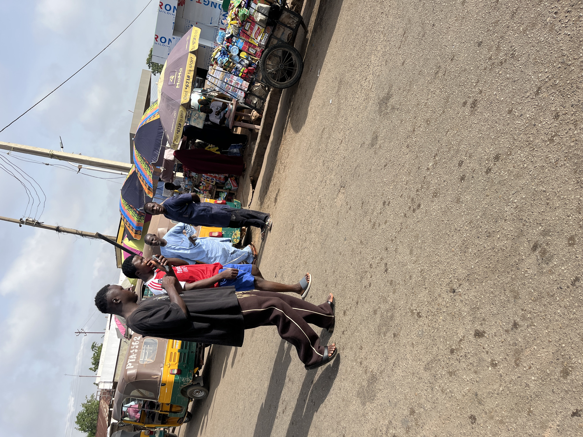 A street scene in Gura Top, Nigeria, features a group of people standing near a vendor's cart laden with goods. An individual in a black shirt and brown pants walks in the foreground, while two other men stand beside him, one in a red shirt and blue shorts, and the other in a light blue traditional robe. Beside them, a man in a dark blue suit stands further back. Parasols advertising "MoMo" and the prompt "Dial *671# to start" are visible above the vending cart. The cart, filled with snacks and beverages, is positioned on the edge of the road. In the background, a "RON BUILDING" sign can be seen. A commercial tricycle vehicle with "PTA-3362" visible on its side is also present. The road surface is cracked and shows signs of wear.