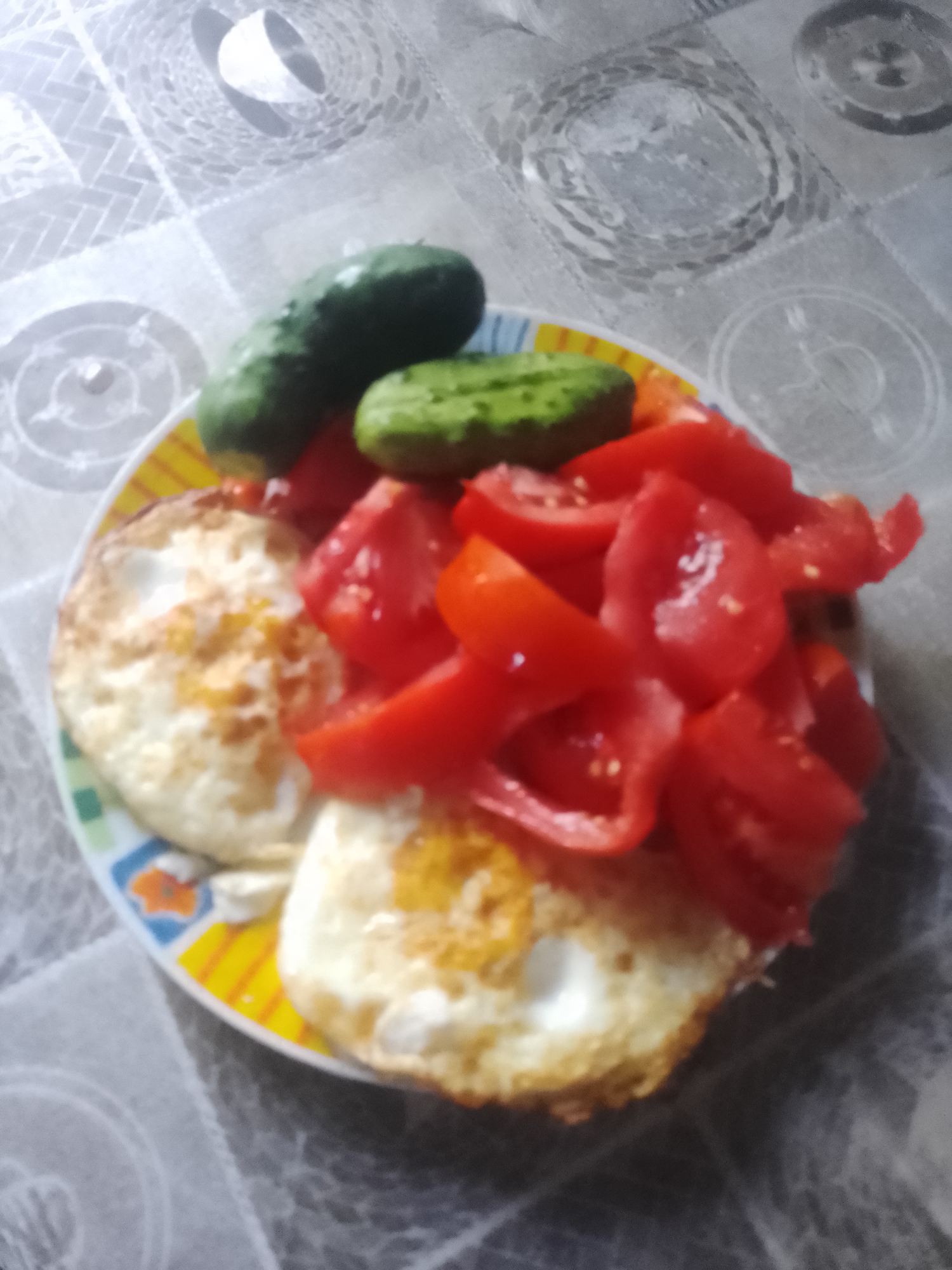 A plate of food, including two fried eggs, sliced tomatoes, and two cucumbers, rests on a tablecloth patterned with grey circles and diamond shapes. The food is arranged on a plate with a colorful border. The scene appears to be indoors, with the tablecloth covering a surface, likely a table. The lighting suggests an indoor setting, possibly a kitchen or dining area. The image is taken from a top-down perspective. The location of this scene within the city of Ustyvytsia, Ukraine cannot be determined from the visual information provided.