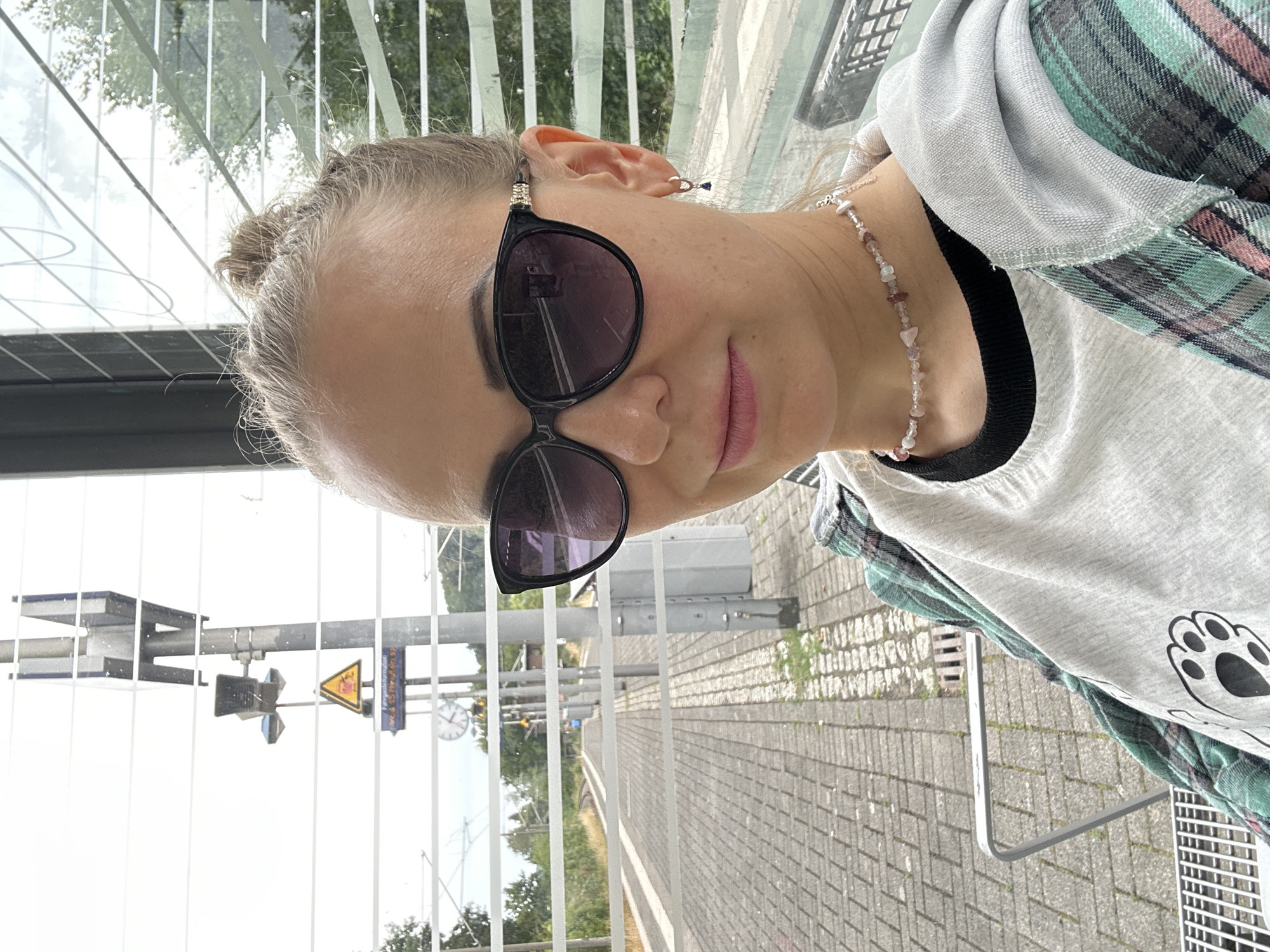 In Velbert, Germany, a young woman with blonde hair tied in a bun sits at a train station platform, evidenced by the tracks and signage. She is wearing large, dark-rimmed sunglasses, a gray t-shirt with a paw print graphic, and a teal and pink plaid shirt over it. A delicate beaded necklace adorns her neck, and a small hoop earring with a dark blue charm is visible in her left ear. The woman is positioned in the foreground, with the train tracks and platform extending into the background. The scene is set within an outdoor shelter, indicated by the slatted glass panels and metal seating. A traffic sign with a yellow triangle and a red symbol is visible on a pole in the background, along with a clock and railway signals. The ground is paved with gray bricks.