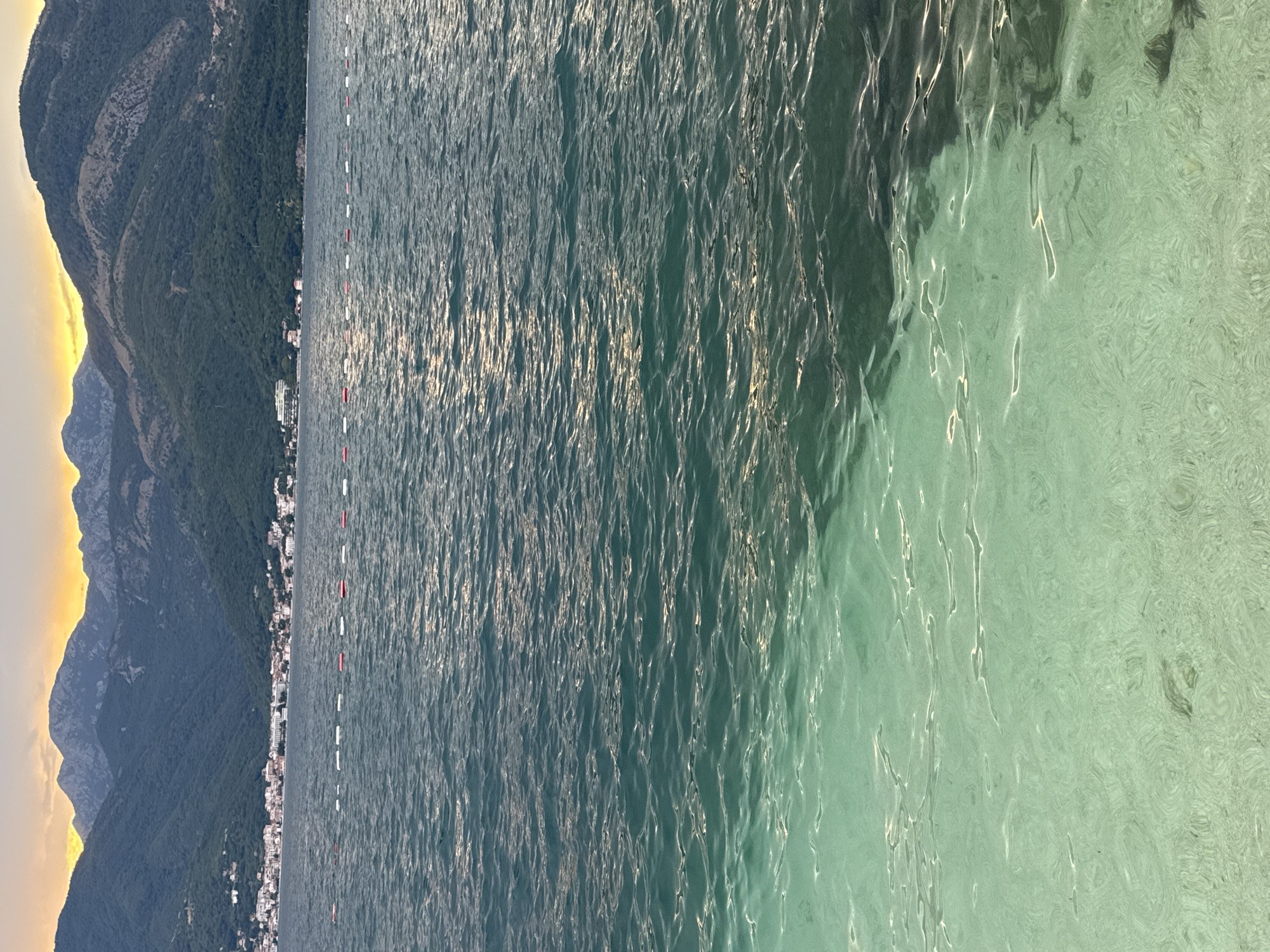 The image is a vertical, wide shot of a body of water with a mountainous coastline in the background. The water is a striking turquoise color, especially in the foreground where it appears shallow and clear, with sunlight reflecting off the rippling surface. Further out, the water deepens to a darker blue-green. A line of buoys, alternating red and white, marks a boundary in the water, extending from the left side of the frame towards the right.
The coastline is characterized by steep, forested mountains that rise dramatically from the water's edge. The vegetation is dense and appears lush green. Nestled along the base of the mountains is a town or village, with buildings visible in shades of white and beige. The slopes of the mountains show evidence of rocky outcrops.
On the far left of the image, the sky is illuminated with the warm hues of either sunrise or sunset, casting a golden glow on the upper portions of the mountains. The light suggests it is either early morning or late evening. The overall mood is tranquil and scenic, highlighting the natural beauty of the location, with the juxtaposition of the imposing mountains and the serene water. No people or specific activities are evident, other than the implied presence of swimmers or boats indicated by the buoys. There is no visible text in the image.