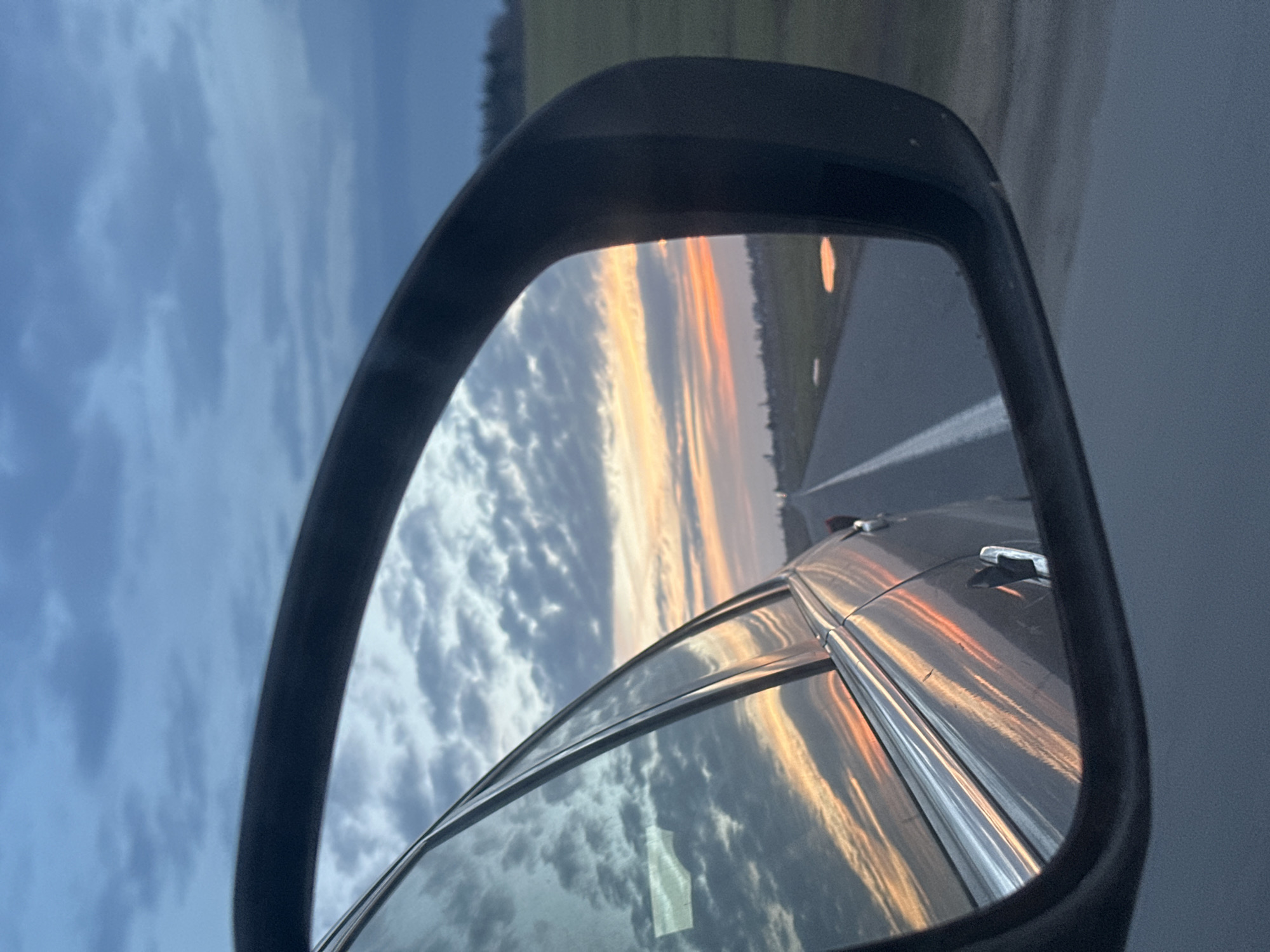 The image displays a view from inside a moving vehicle, focused on the passenger-side rearview mirror and a portion of the vehicle's exterior. The foreground on the left shows the reflective surface of the car's window and part of its grey body, reflecting blue and orange cloud formations. The prominent object is a black-framed side mirror, reflecting a scene consistent with a late evening or early morning sky over a rural road in Liozna, Belarus.

Within the mirror's reflection, a straight asphalt road extends into the distance, marked by a solid white line on its right edge. Adjacent to the road, a green field is visible, containing several shallow puddles of water that reflect the sky's colors. The horizon shows a blend of dark blue and grey clouds transitioning to vibrant orange and pink hues near the setting or rising sun, indicating twilight conditions. A faint treeline is discernible in the far distance along the horizon. No individuals are visible. The car's door handle, in a metallic finish, is partially visible within the mirror's reflection, suggesting a grey or silver vehicle.