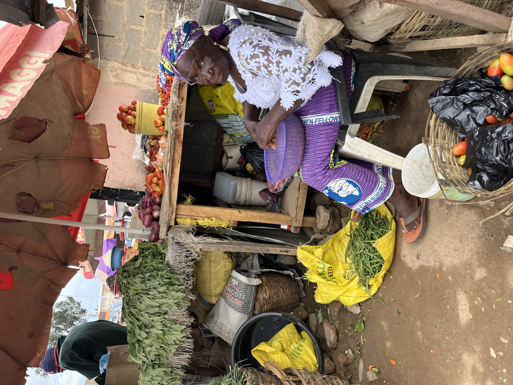 This image depicts an outdoor market stall in Jos, Nigeria. Two individuals are present amidst various produce.
In the foreground, on the right, an elderly dark-skinned woman is seated on a dark plastic chair. She wears a patterned headwrap with shades of purple, yellow, and blue, a white lace top with an embossed floral pattern, and purple trousers featuring white text, partially legible as "MEN-FELLOWA." She holds a purple plastic sieve or bowl, appearing to prepare orange-colored vegetables, fragments of which are scattered on the dirt ground. Gold-colored earrings are visible, and she looks directly at the camera. Her feet wear orange and brown flip-flops.
To the left, another individual is seen from behind, wearing a dark green long-sleeved top and a blue and red knitted beanie. They stand behind a wooden display stand.
The market stall features multiple piles of produce. The left display stand holds large bunches of green leafy vegetables with visible roots. The right display stand contains piles of red onions, a yellow plastic bucket filled with red and orange tomatoes, and a separate mound of small red peppers.
The ground is uneven, packed dirt, scattered with small rocks and vegetable debris. A yellow plastic sack, partially marked "KIKURU FEED," rests on the ground near the seated woman's feet, containing a heap of green string beans. A large woven basket in the immediate foreground holds red and orange tomatoes, along with a black plastic bag. Additional woven baskets and yellow sacks are visible on the ground and beneath the display stands. A white sack, partially labeled "ANGOTE MASTER," is propped against a wooden support structure in the background.
The stall is covered by a large, tattered reddish-brown market umbrella, with "MAGGI" branding partially visible. Concrete block walls are visible in the background, with an overcast sky above.