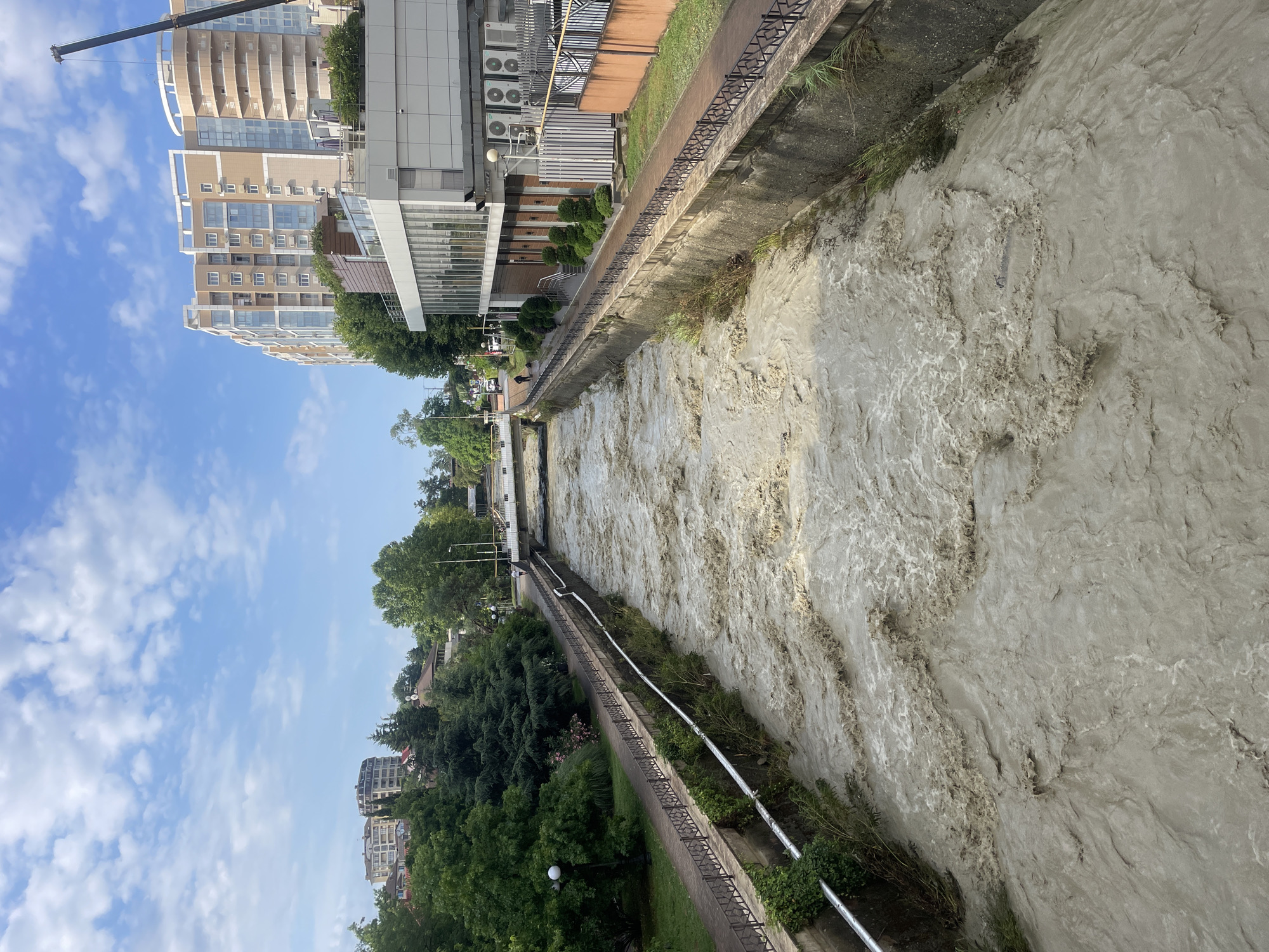 The image depicts a wide, fast-flowing river with murky, brown water carrying debris. The river is channeled between concrete embankments lined with metal railings. On the left bank, a paved pathway runs alongside a grassy park with trees and street lamps. On the right bank, there is a paved walkway adjacent to a modern, multi-story building with large windows and balconies, and further along, another building with an orange facade and outdoor seating. The sky above is blue with scattered white clouds. In the distance, several buildings of varying heights and architectural styles are visible, along with more trees. The scene suggests a city environment, likely in Sochi, Russia, given the context. The turbulent water indicates recent heavy rainfall or flooding. There are no visible people in the immediate foreground of the river, but a single person is visible walking on the pathway on the right bank near the building.