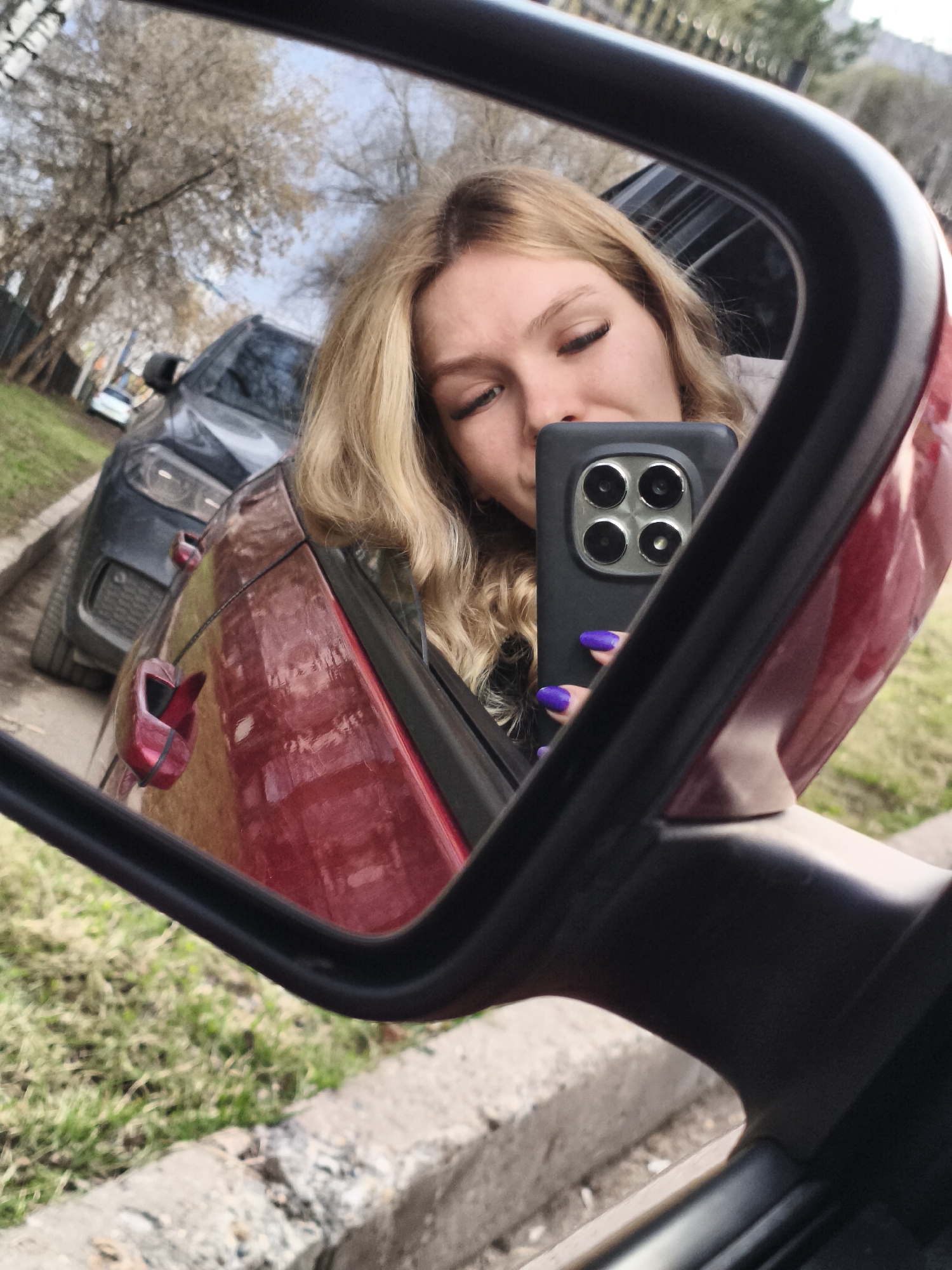 A selfie is being taken in the side mirror of a red car. The reflection in the mirror shows a woman with blonde, wavy hair and purple fingernails, holding a black smartphone. The mirror's surroundings show the red car door, the road, green grass, a gray curb, and a gray SUV parked nearby. The background also shows trees with bare branches. The scene is located in Izhevsk, Russia.
