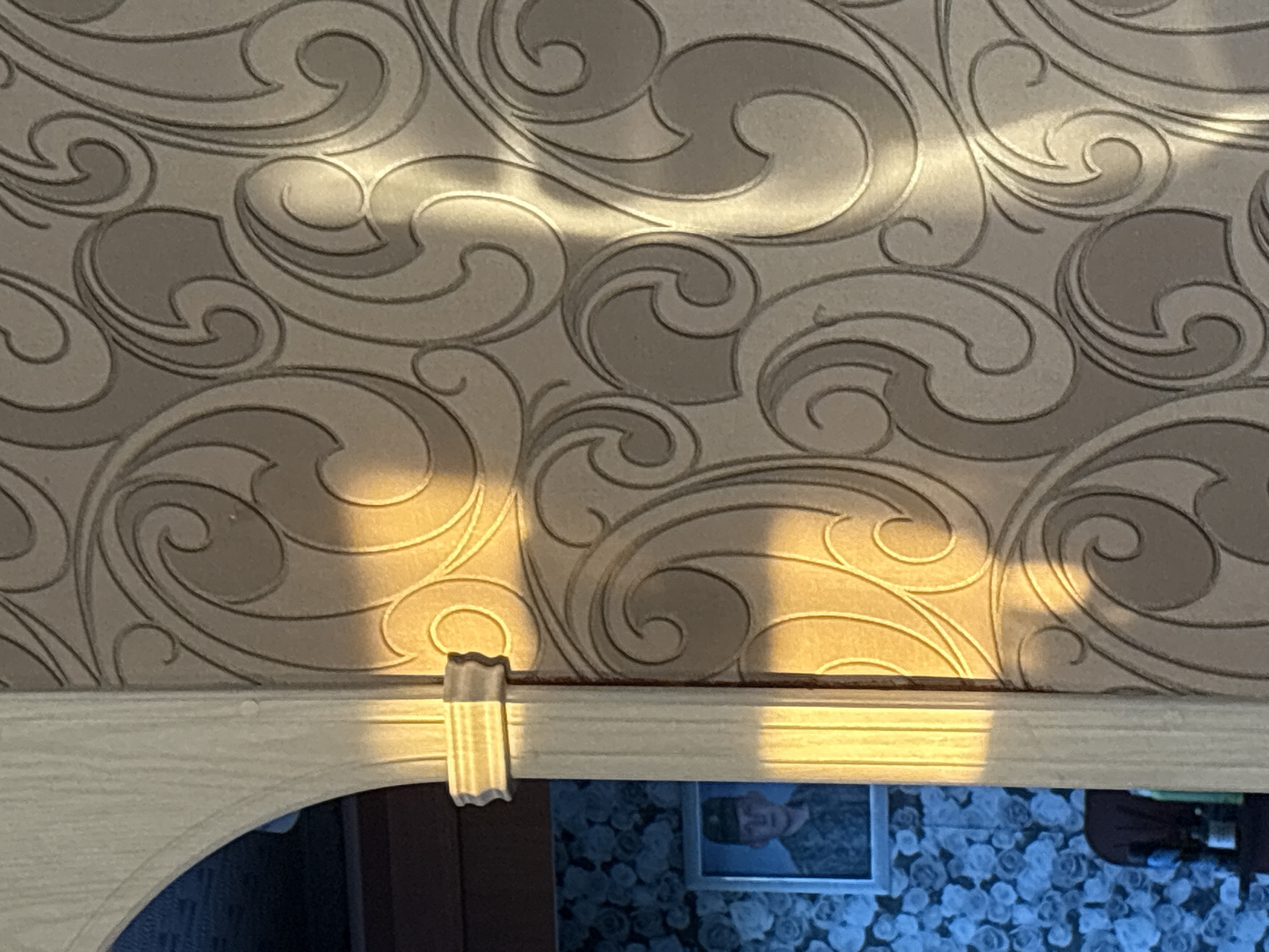 The image depicts a room with patterned wallpaper. A textured, off-white architectural element, possibly a column or archway, is visible on the left side, casting a shadow. Sunlight strikes this element, creating a warm glow. Partially visible through an opening in this structure is a framed portrait of a man in a military uniform and a dark cap. He is looking directly at the camera. The background behind the portrait features blue wallpaper with a floral pattern. The overall impression is of an interior domestic setting. The location is identified as Sovkhoza "Razdol'e", Russia.