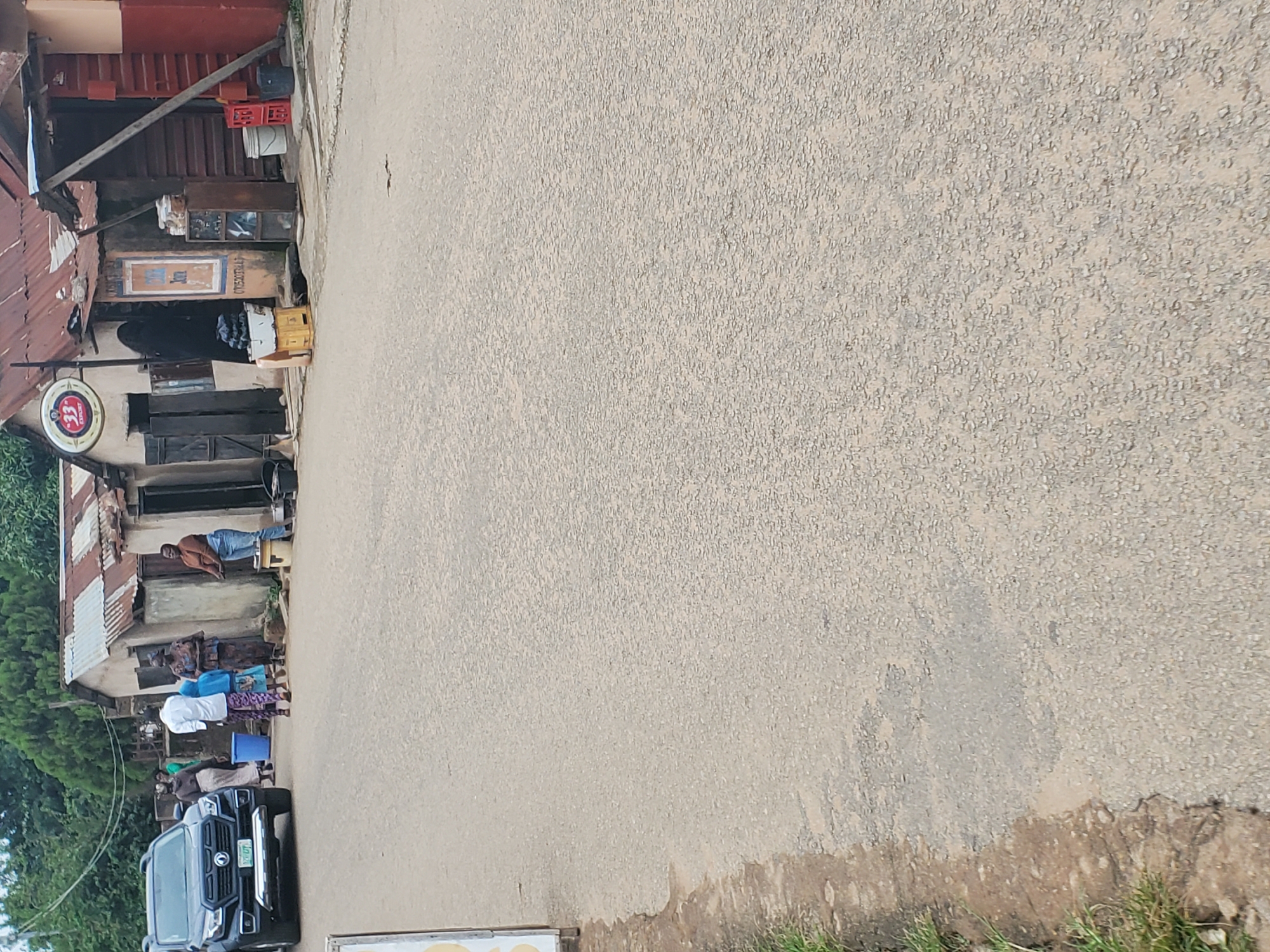 The image depicts an outdoor scene in Jos, Nigeria, likely in a rural or semi-urban area. A roadside structure, possibly a shop or bar, is visible on the left, with a corrugated iron roof and weathered walls. In front of the structure, several people are gathered, some standing and others seated.
A man in a brown jacket and blue jeans stands near a doorway, looking towards the camera. Several women in colorful traditional attire are also present, suggesting a social gathering or community event. A dark-colored SUV is parked in the foreground, facing the building.
Notable details include several crates stacked near the entrance of the building, some appearing to be for beverages. A circular sign with the number "33" and text below is hanging from the structure, likely indicating a brand or business name. The ground is a paved road, showing signs of wear and tear. The vegetation on the far left suggests a green, possibly tropical environment. The lighting indicates it is daytime, and the weather appears to be clear. The overall impression is one of everyday life and community interaction in a Nigerian setting.