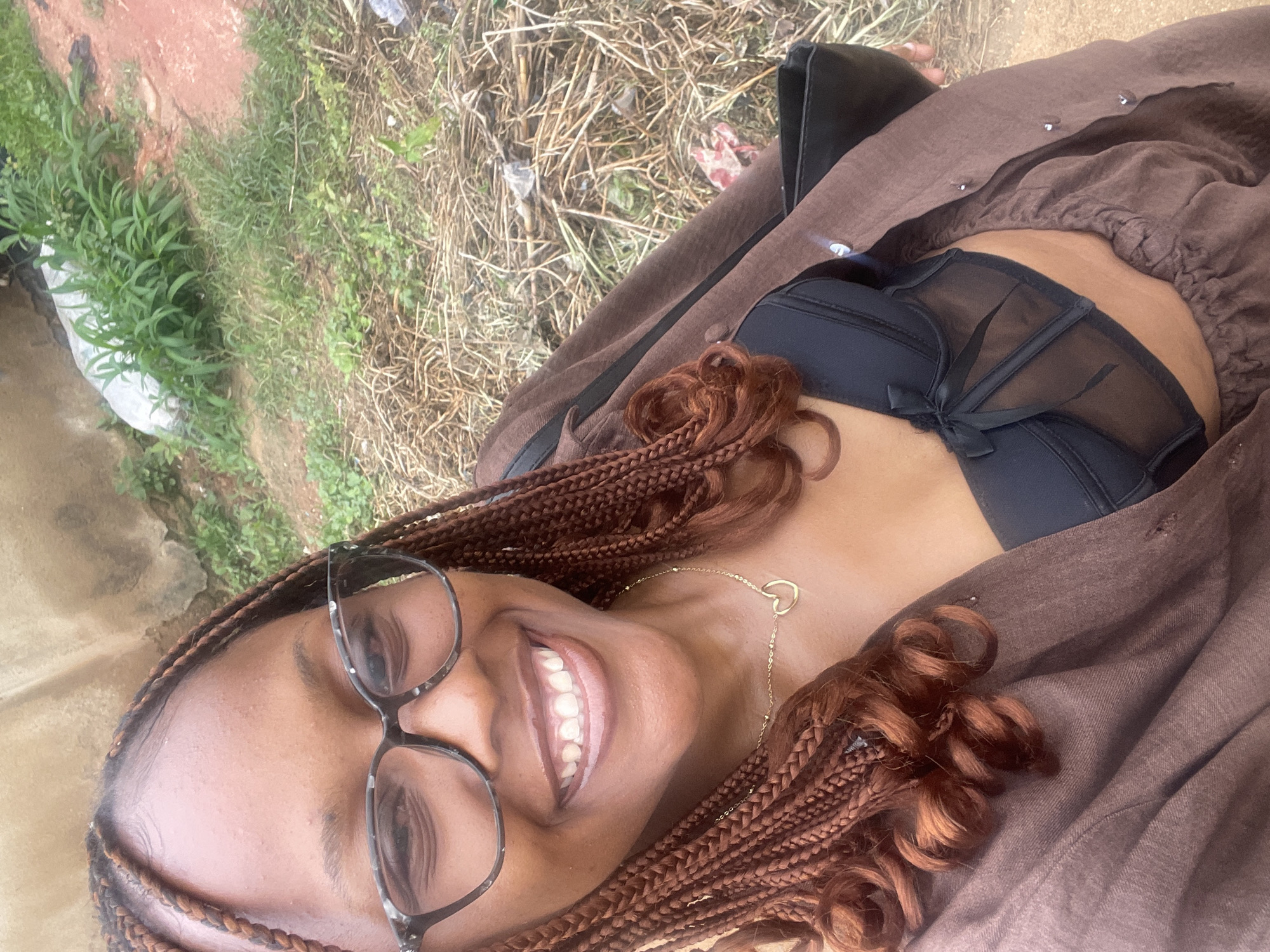 A woman with braided brown hair and glasses smiles at the camera. She is wearing a black bra with sheer paneling and a brown open jacket. A gold heart-shaped necklace is visible on her chest. She is holding a black purse in her right hand. The background shows dry grass and foliage, suggesting an outdoor environment. The image is taken at a medium-close range, focusing on the woman's upper body. The location is identified as Rafin Par, Nigeria.