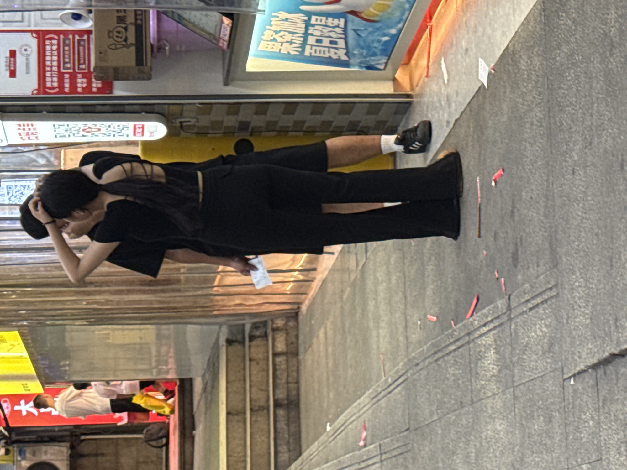A young Asian woman and a young Asian man, dressed in black, are standing on a paved sidewalk in front of a building with a reflective, corrugated metal facade. The woman, facing away from the camera and towards the man, has her hair pulled back in a ponytail and is touching the back of her head with one hand. She is wearing a black sleeveless top with a criss-cross detail on the back and black flared pants. The man, partially obscured, is facing the woman and has his arm around her shoulders, also touching the back of her head. He is wearing a black t-shirt and black shorts. A small white slip of paper is held in the man's right hand. The sidewalk is littered with small red and white pieces of paper. In the background, a person in a white t-shirt and dark pants, holding a yellow bag, walks up a set of stairs. The building facade has various signs, including one with red characters and a QR code. A display case with blue and white text and images is to the right of the subjects. The scene appears to be outdoors in an urban environment in the city of Yuzhong District, China.