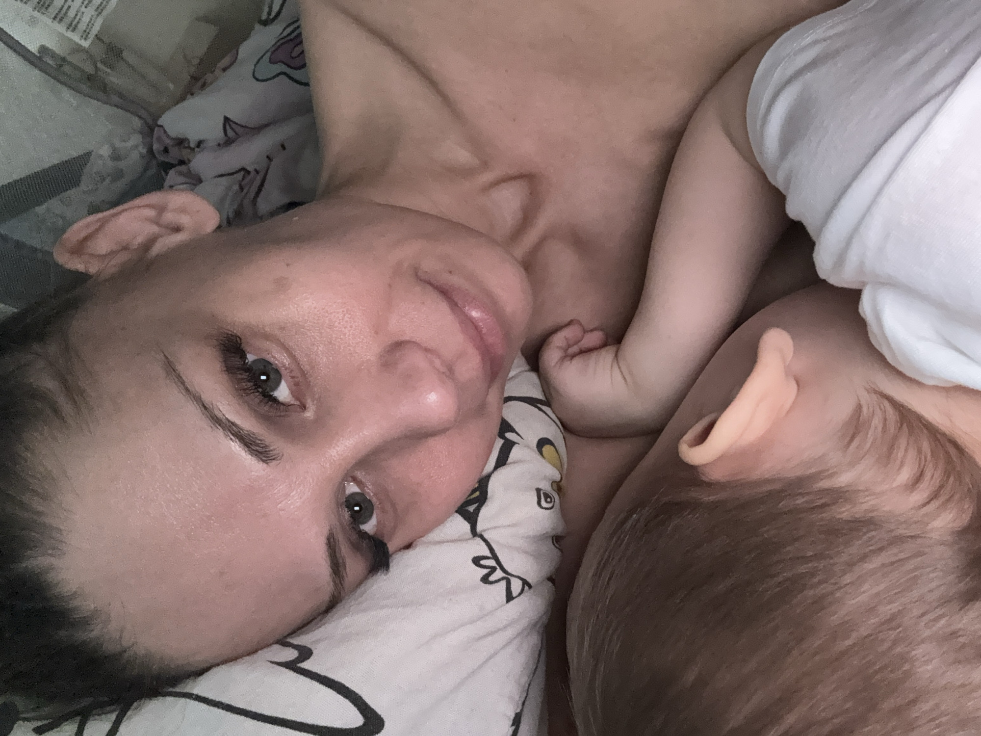 A close-up, slightly angled shot shows a woman lying in bed next to a baby. The woman is fair-skinned with dark hair pulled back, and she is looking at the camera with a slight smile. The baby, presumably sleeping, is partially visible with brown hair and a white garment. The bedding is patterned with black and white designs. The image does not provide any evidence to indicate the location in the city of Zhukovo, Russia.