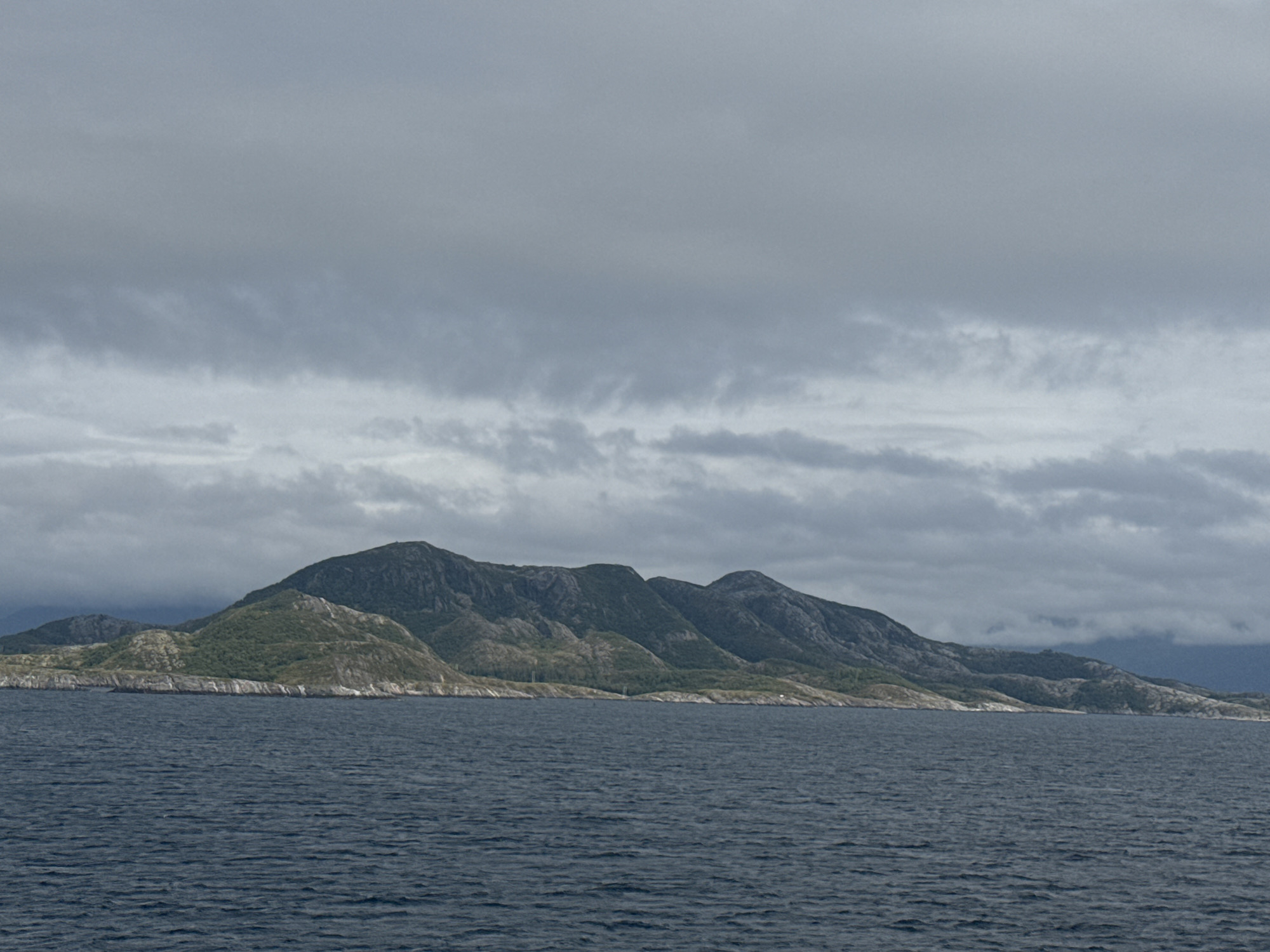 The image depicts a scenic view of the island of Onøya, Norway. In the foreground, a body of dark blue water stretches across the frame, with a subtle ripple texture indicating a calm surface. The midground is dominated by the island's mountainous terrain. The slopes are covered with patches of green vegetation and rocky outcrops, suggesting a rugged landscape. The mountains are varied in height and shape, forming a cohesive ridge that extends horizontally across the image. In the background, a cloudy sky fills the upper portion of the frame. The clouds are predominantly gray and appear to be layered, with some darker, more dense formations and lighter, wispy sections. The overall impression is a cool, overcast day in a coastal mountainous region. There are no people or man-made structures clearly visible in this image, focusing solely on the natural environment.