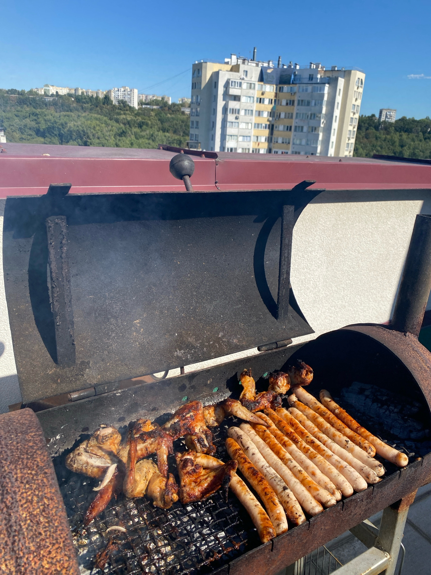 An outdoor barbeque is in progress on a rooftop with a cityscape in the background. The barbeque, a rusty barrel-style grill, contains chicken wings and several rows of sausages cooking on the grate. Smoke rises from the grill, indicating that the food is actively being cooked. The background shows a clear blue sky, lush green trees, and a cluster of multi-story apartment buildings. The buildings are typical of Soviet-era architecture, common in many Eastern European cities, including Chișinău, Moldova. The scene suggests a warm, sunny day, perfect for an outdoor meal.