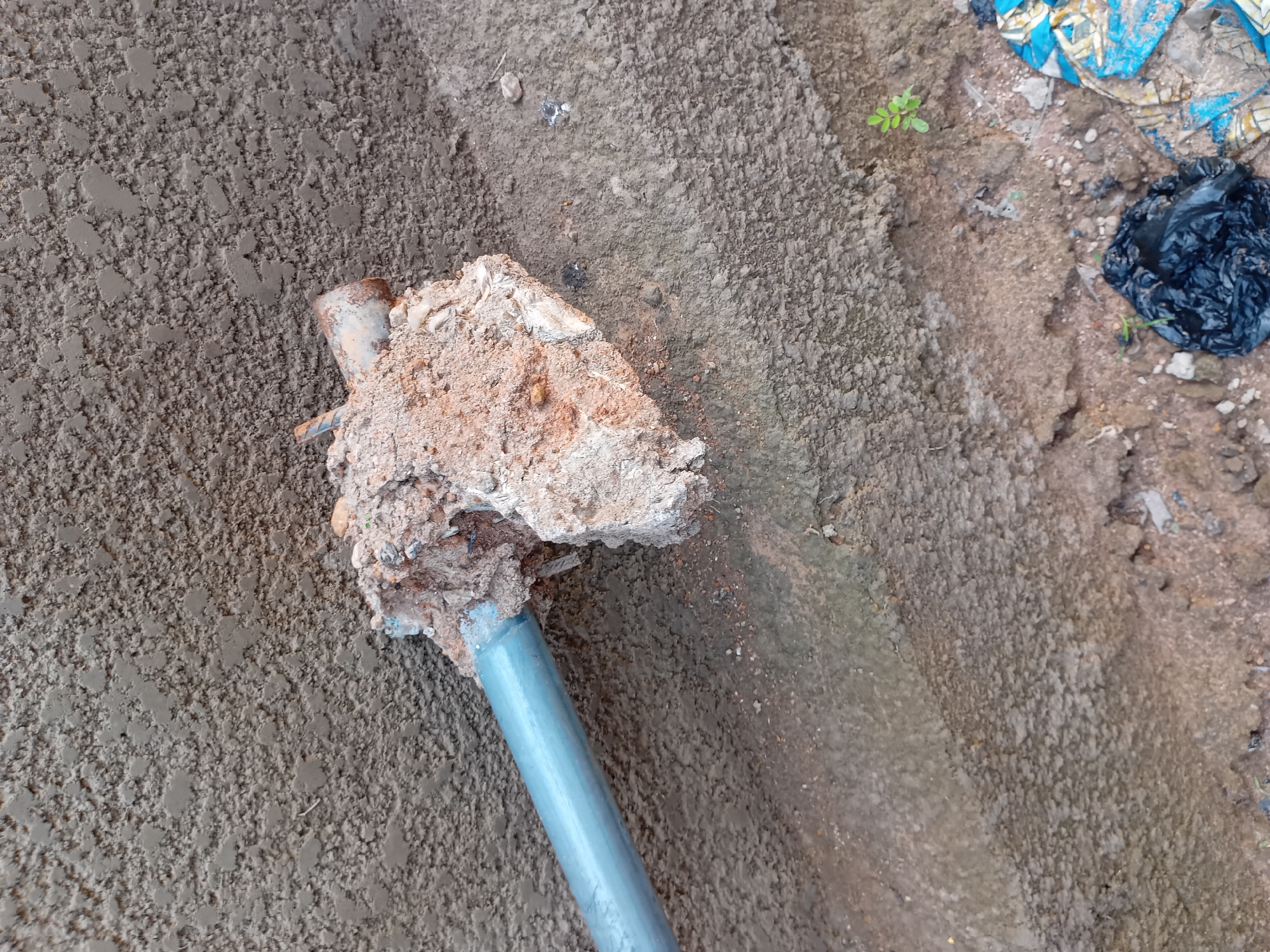 The image shows a close-up view of a metal pipe protruding from a textured, grey concrete wall. The pipe appears to be partially embedded in a clod of dried dirt and concrete debris, which is also attached to the wall. The surrounding environment is rough and appears to be outdoors, with dirt and loose stones visible at the base of the wall. There is also some litter, including a black plastic bag and a piece of blue and yellow patterned material, on the ground. The overall impression is of an unfinished or neglected outdoor area, possibly a construction site or a backyard in Jauro Yinu, Nigeria.