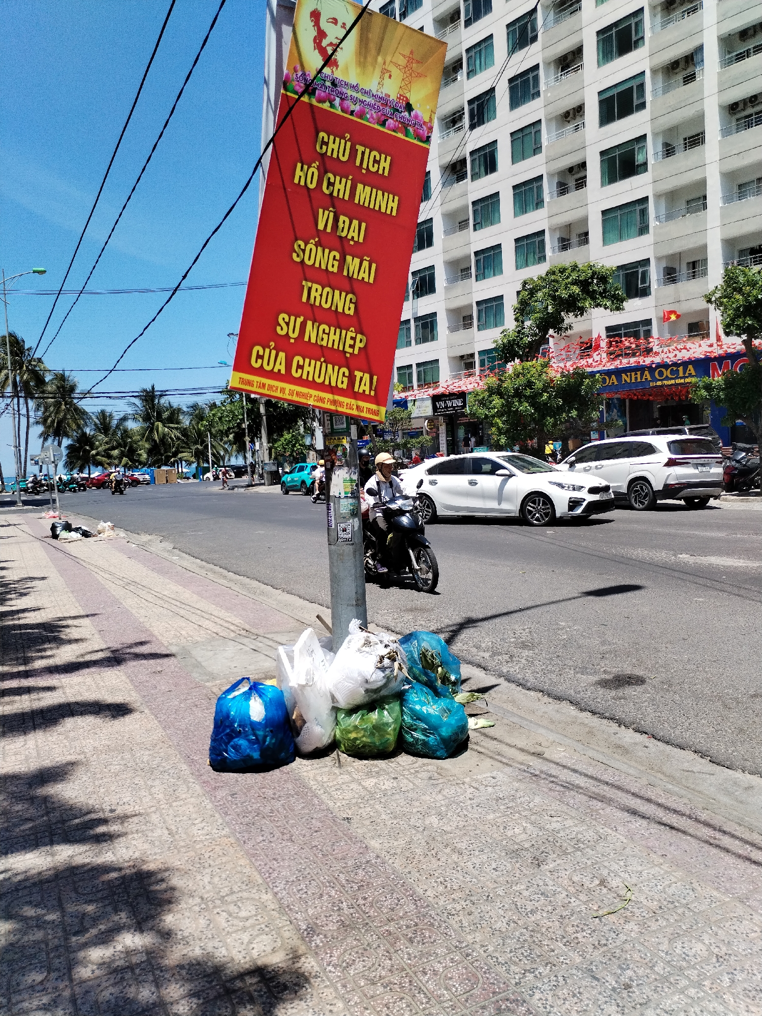 The image captures a scene on a sunny day in Bac Nha Trang Ward, Vietnam, specifically on a street bordered by a sidewalk and a building facade. The sidewalk is paved with patterned tiles and strewn with shadows from nearby trees. Several full trash bags, colored blue, white, and green, are piled up near the curb, close to a metal pole with a large red banner attached. The banner features an image and text in Vietnamese, including the words "CHU TICH HO CHI MINH" (Chairman Ho Chi Minh).
A motorcyclist wearing a hat and dark clothing is riding a black motorbike in the street, moving from left to right, away from the trash and banner. Two white vehicles are visible further down the street, a sedan and a larger SUV, along with other vehicles in the distance.
In the background, across the street, a multistory white building is observed with numerous windows and balconies. The building's facade is adorned with red decorations, which appear to be flags or banners. The street is lined with palm trees and other greenery, adding to the tropical setting. Overhead, several power lines stretch across the clear blue sky.