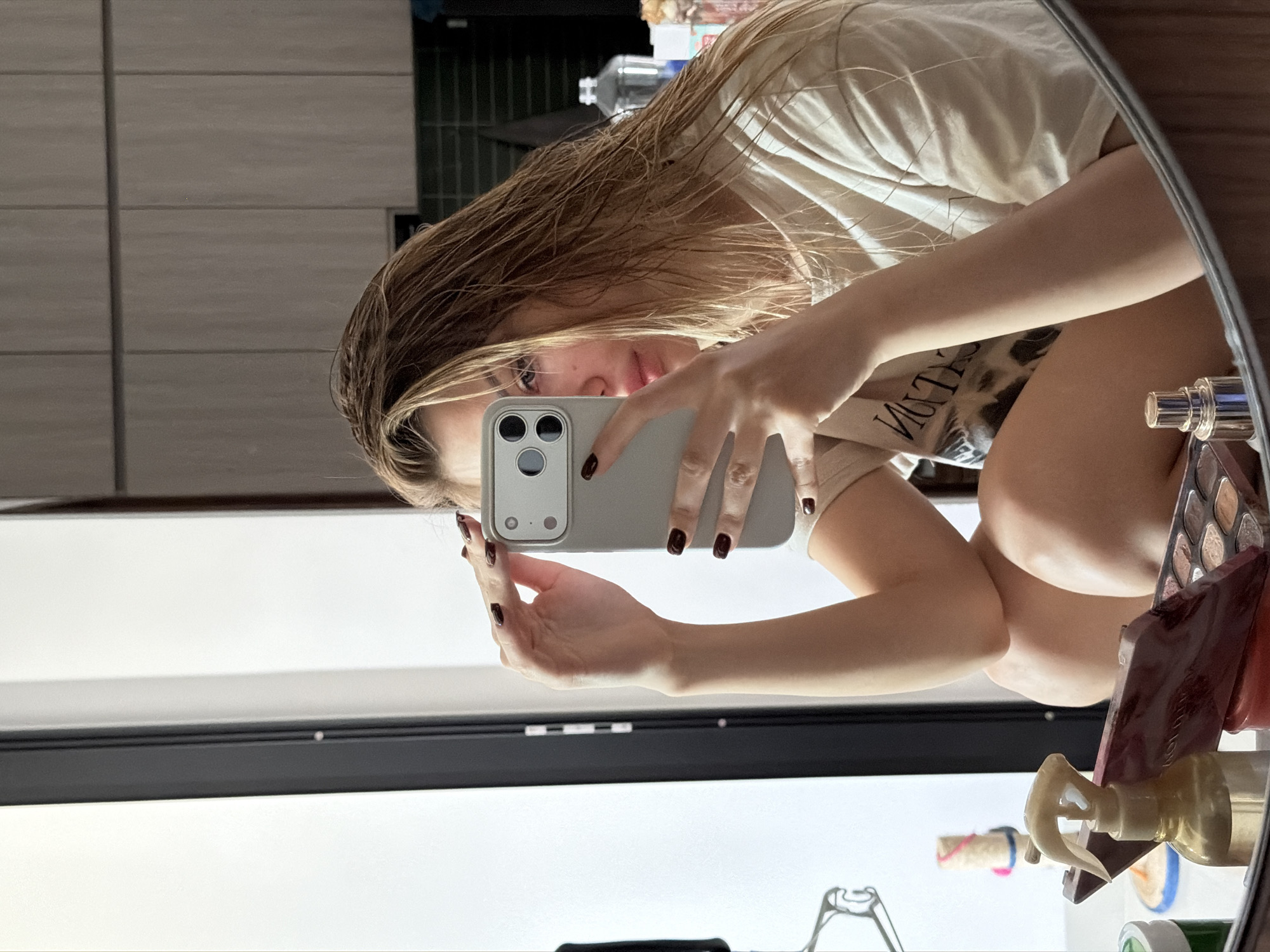 A young woman with wet, blonde hair takes a selfie in a bathroom mirror. She is wearing a light-colored t-shirt. Her left hand is holding a smartphone, and her right hand is touching her hair. In the foreground, a makeup palette and a spray bottle are visible on a counter. The background shows kitchen cabinets and a large window. The image does not contain any identifiable landmarks or elements that would suggest a specific location in Da Nang, Vietnam.