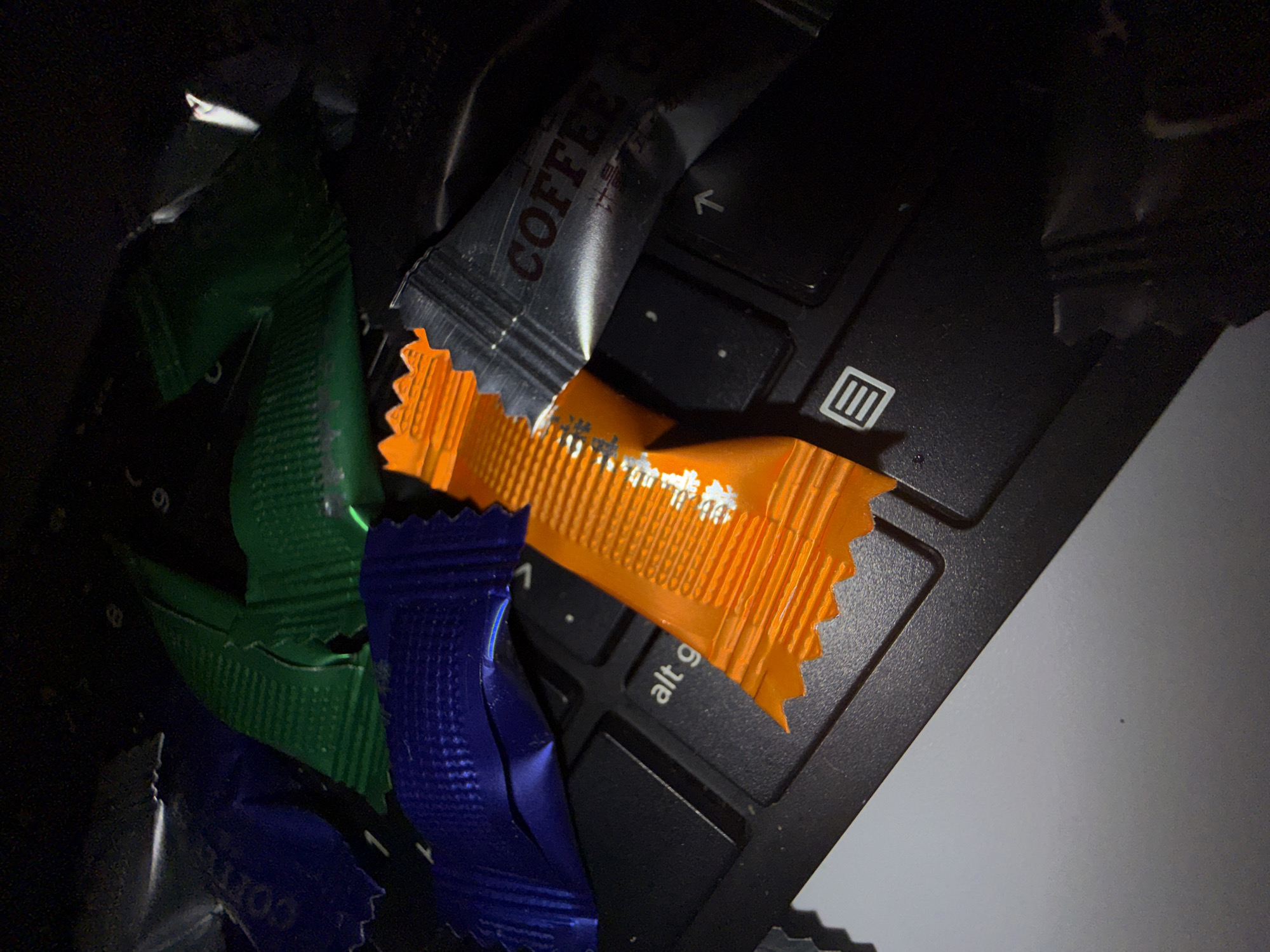 The image shows wrappers from coffee creamer packets scattered on a black laptop keyboard. The wrappers are various colors including green, orange, blue and silver. The word "COFFEE" is visible on one silver packet. The context of location in Lupanovo, Russia is not discernible from the visual information provided.