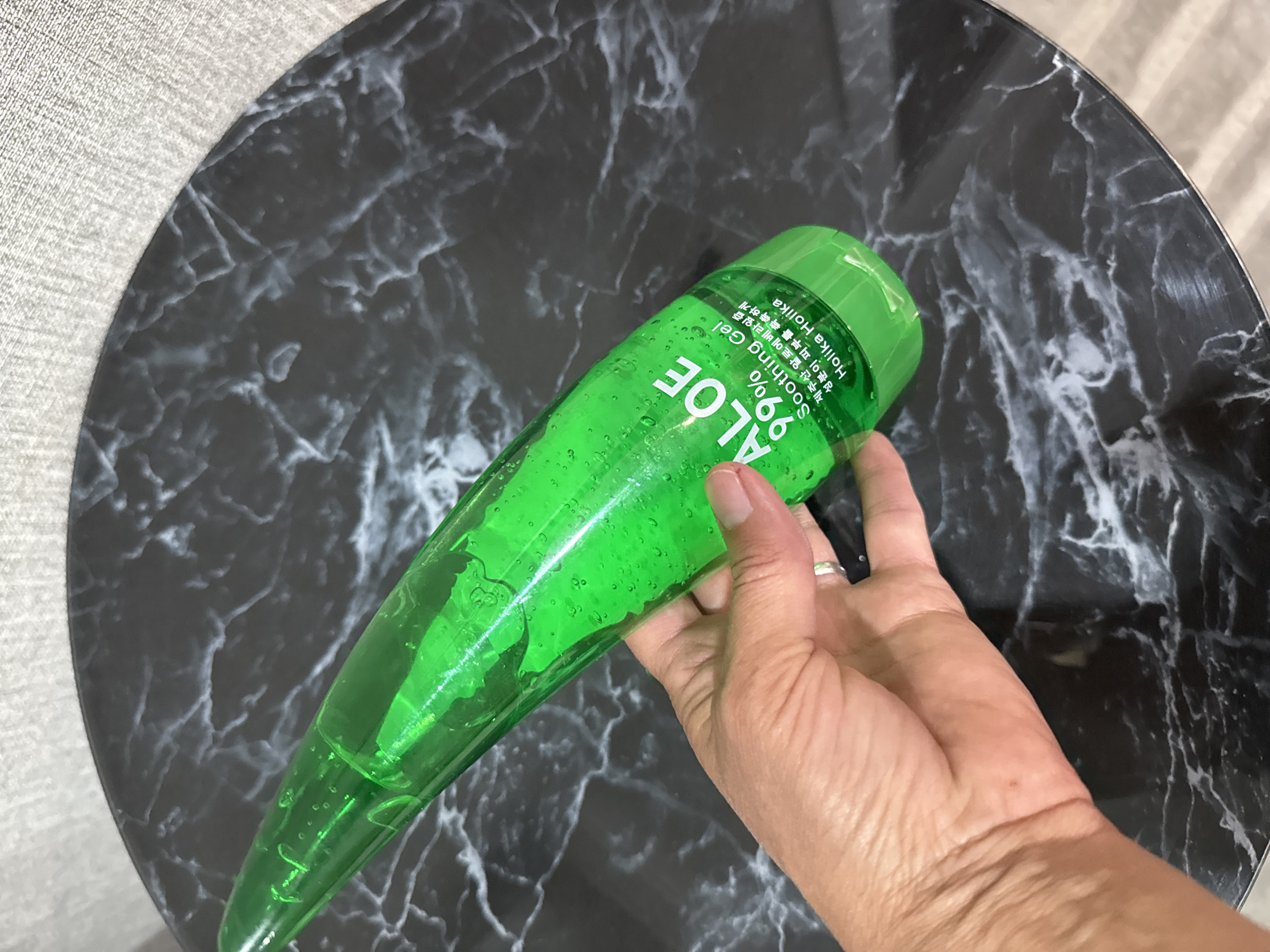 A hand holds a clear green plastic bottle of "ALOE 99% Soothing Gel" which is placed on a round black marble-patterned table. The bottle is angled diagonally across the image, with the top pointing towards the upper left. The hand is on the left side of the image, gripping the middle section of the bottle. The table has a textured, light-colored background behind it. The scene does not provide any information about the city of Солонешное, Russia.