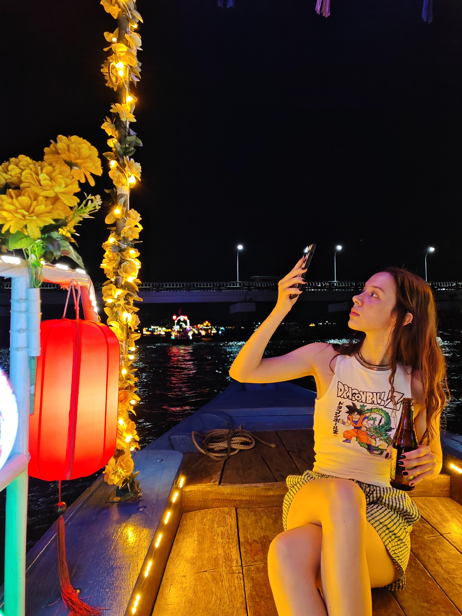 This image captures a nighttime scene on a boat along the Thu Bon River in Hoi An, Vietnam, a city known for its lantern-lit ancient town and vibrant river activities. The boat is adorned with yellow flowers and string lights that illuminate the wooden deck and a large red lantern. In the foreground, a young woman with long brown hair is seated on the boat. She is wearing a white tank top featuring a Dragon Ball Z graphic and a striped skirt. The woman is holding a smartphone in her right hand, seemingly taking a photo or video, and a brown bottle in her left hand. The background shows the dark river with reflections of distant lights, a bridge, and other boats, suggesting a festive atmosphere. The overall lighting is dim, with the boat's decorations and the distant city lights providing the primary illumination.
