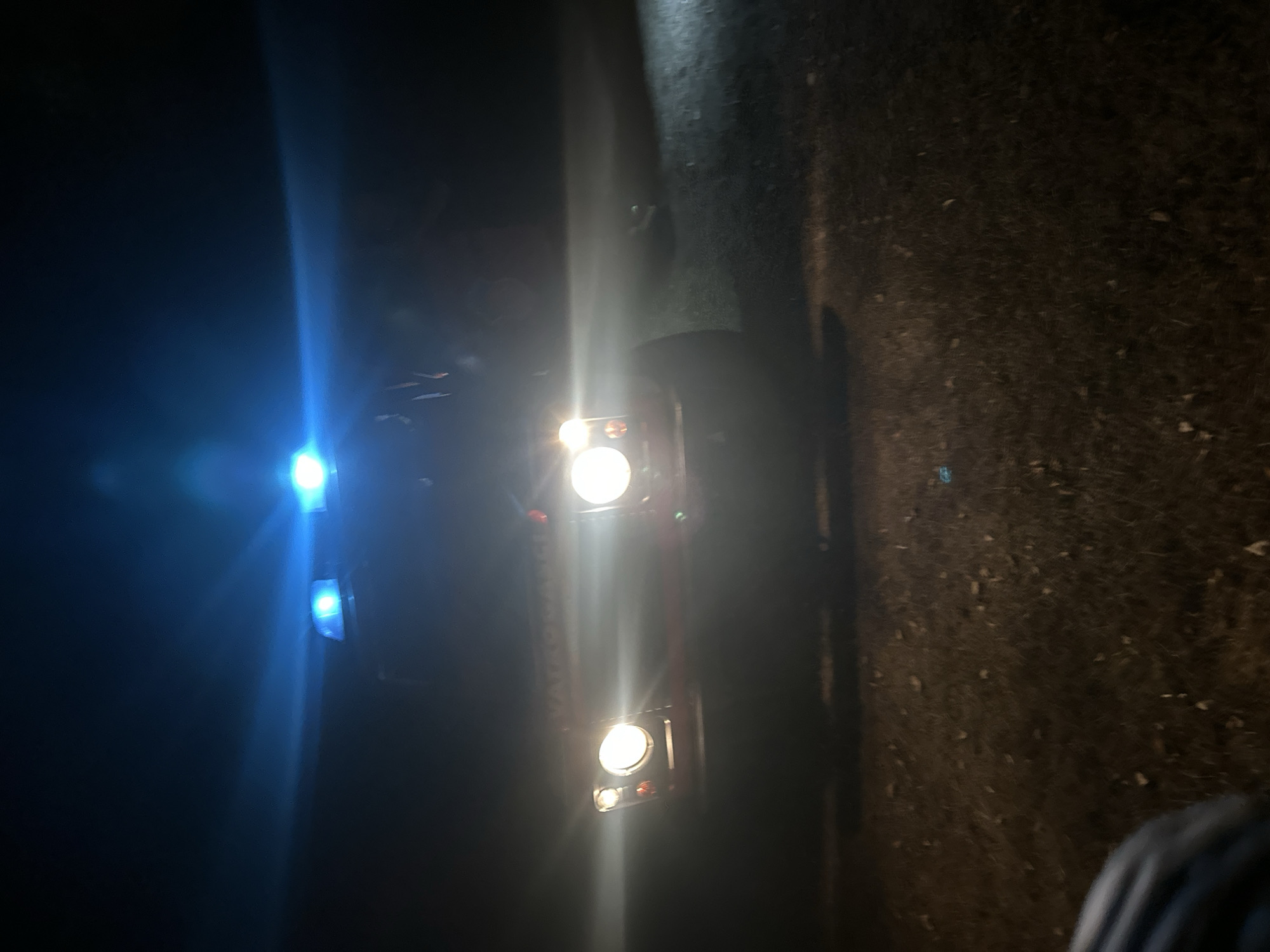 An unflared, close-up, low-angle, medium shot captures the front of a red vehicle at night. The vehicle has its headlights on, emitting bright white light that overpowers much of the front of the vehicle. Above the headlights, two blue emergency lights are illuminated, casting a bright blue glow upwards and outwards. The vehicle appears to be a utility vehicle or off-road vehicle, possibly a Land Rover Defender, given its boxy shape and prominent headlights. A portion of what appears to be a tire is visible in the lower left of the frame. The ground beneath and in front of the vehicle is dark and appears to be covered in grass or low vegetation with scattered leaves. The background is entirely dark, indicating a nighttime scene in an outdoor environment, potentially a rural area. The location is identified as Bijele Poljane, Montenegro.