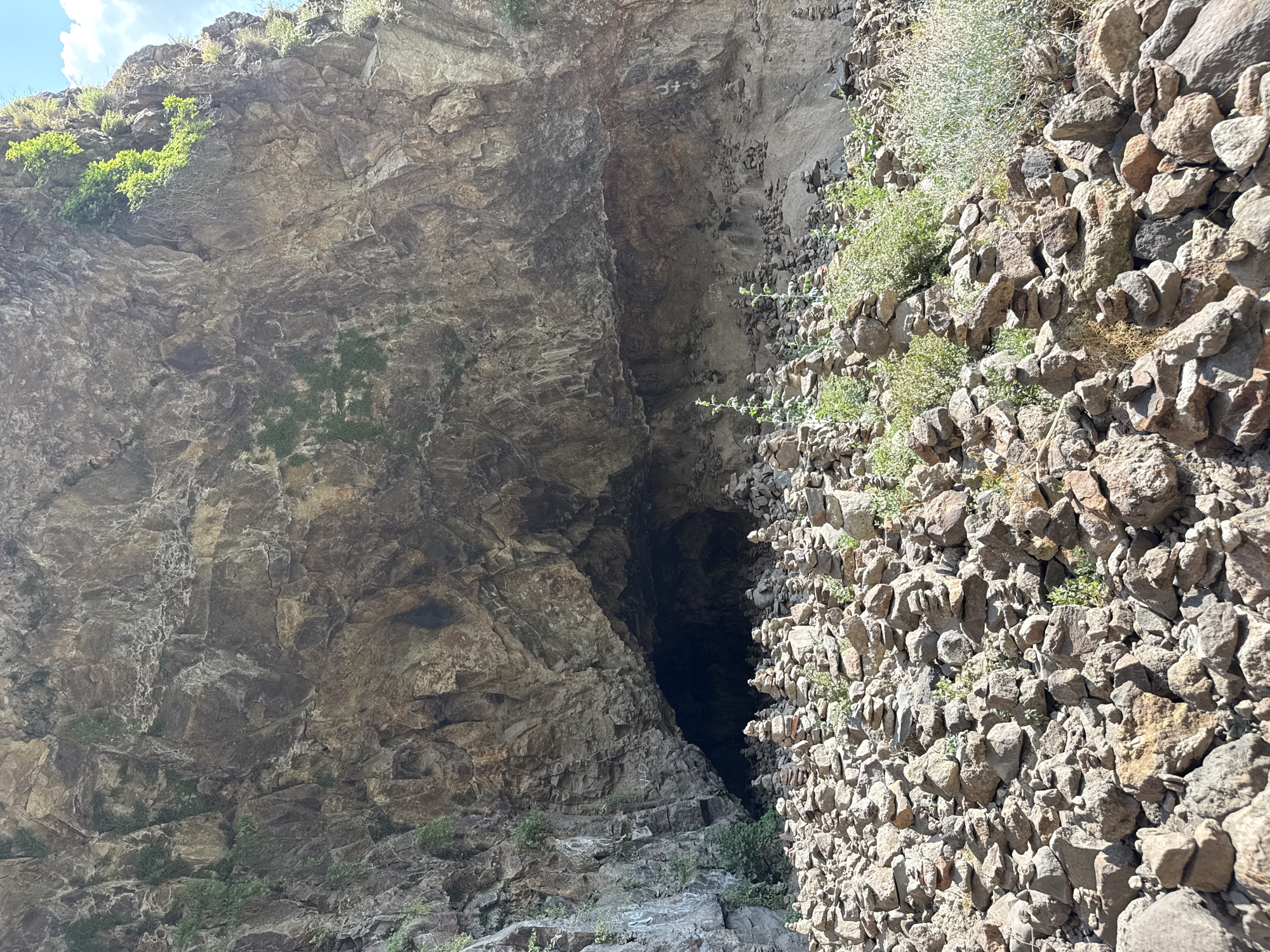 The image depicts a natural scene at the Devil's Bridge (Satan's Bridge) in Goght, Armenia, a geological formation characterized by a large cave-like overhang carved into a rocky cliff face. The foreground is densely populated with numerous stacked rock formations, known as cairns, varying in size and height, suggesting a place of human activity or tradition. Some of these cairns are quite tall, while others are smaller and more haphazardly arranged. Green vegetation, including grasses and bushes, grows sporadically amongst and behind the rock piles, indicating a natural, outdoor environment. The cliff face itself is rough and textured, with layers of rock and occasional patches of greenery clinging to its surface. The entrance to the cave is visible in the mid-ground, appearing dark and recessed into the cliff. The overall lighting suggests daylight, with shadows cast by the rocky overhang and the cairns. The sky is partially visible at the top right of the image, with scattered clouds. There are no people or animals directly visible in the scene.