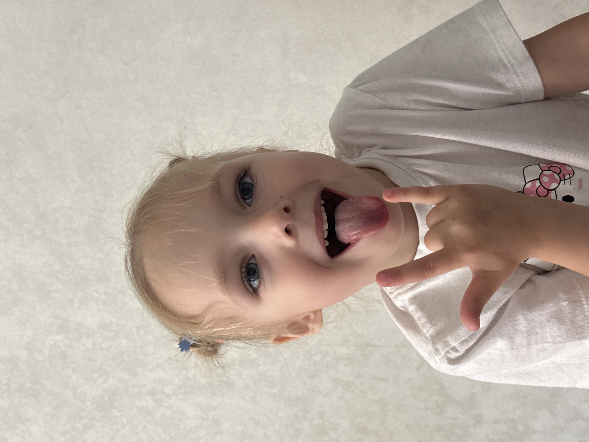 A young, light-skinned child with blonde hair pulled back in a small ponytail with a blue hair tie, is sticking her tongue out and making a hand gesture. She is wearing a white t-shirt with a print of a Hello Kitty design. The background is a textured off-white surface. The image is taken at an upward angle. There is no evidence to confirm that the image was taken in the city of Chaikovsky, Russia.