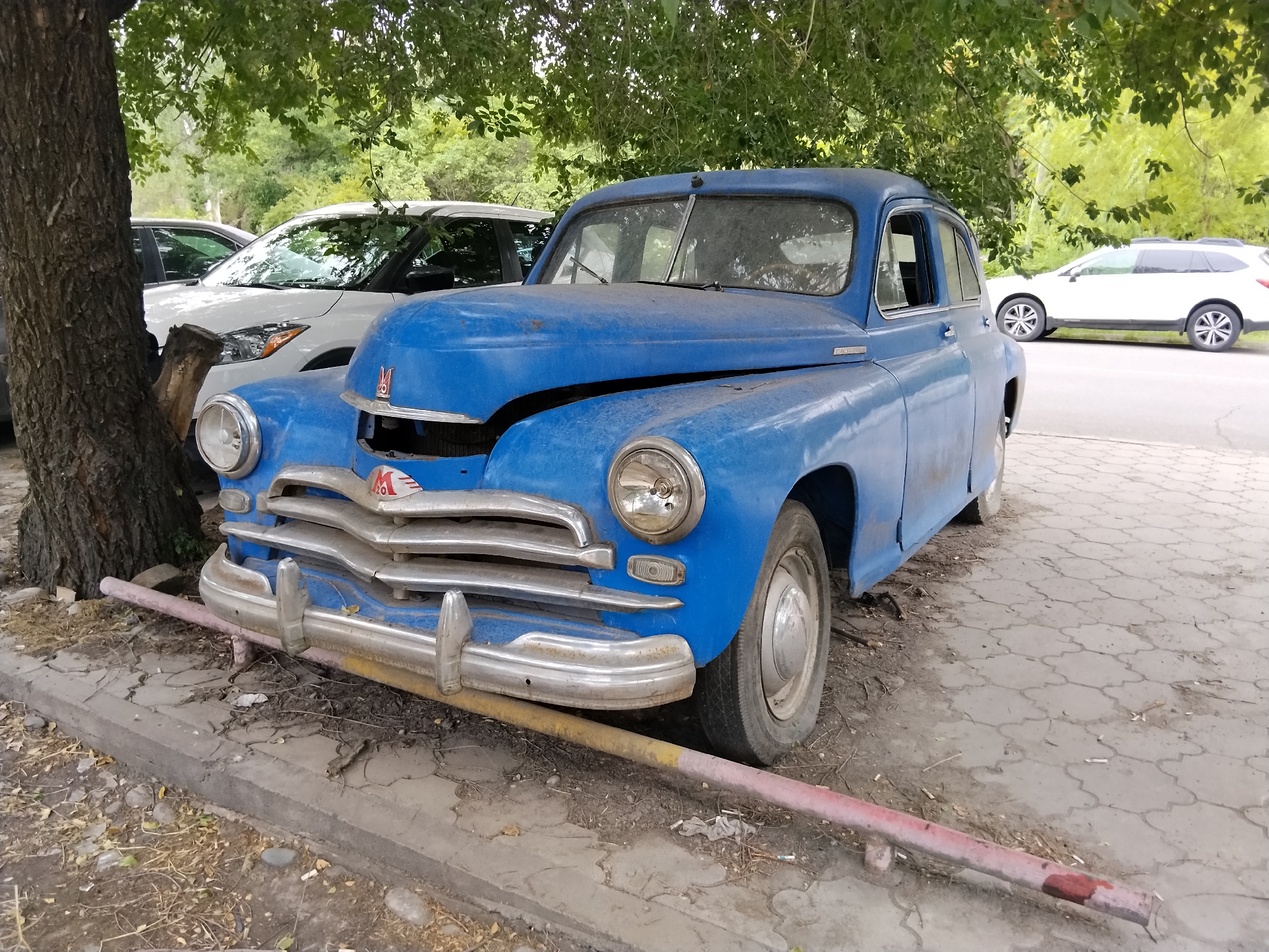 In Bishkek, Kyrgyzstan, a blue, vintage GAZ car is parked on a cracked concrete sidewalk under a leafy tree. The car is dusty, with some rust visible. Its hood is slightly ajar, revealing part of the engine. The car has chrome accents on the grill and bumper. A white SUV is parked behind it. The environment appears to be an urban area, possibly a residential street or a park.