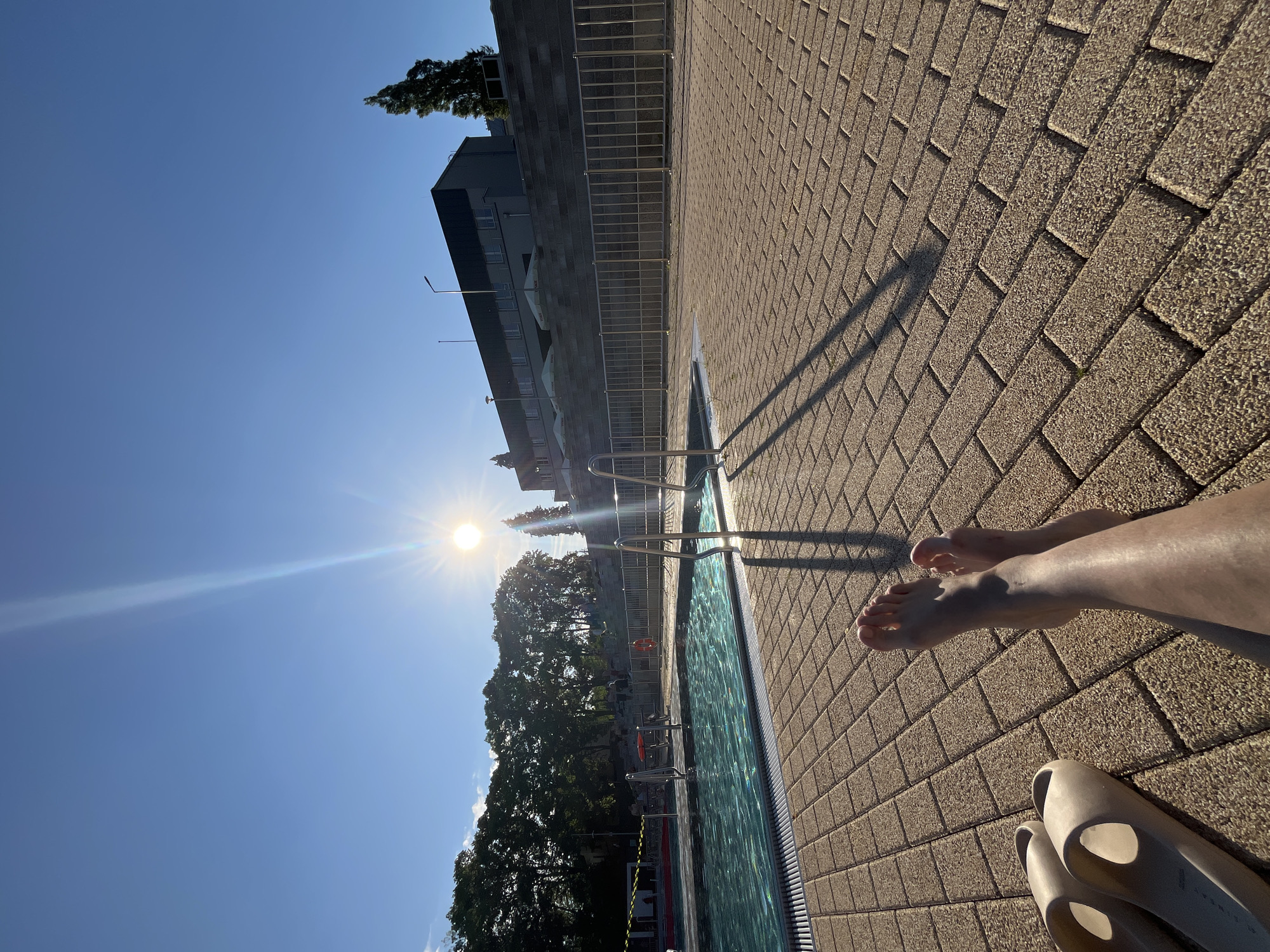 The image depicts an outdoor swimming pool area under a clear, bright blue sky with a low sun causing significant lens flare and long shadows. In the immediate foreground, light-skinned bare legs and feet are visible, resting on a textured paved surface. To the lower left, a pair of light beige slip-on sandals are positioned. The paving consists of rectangular, light brown pavers with a visible granular texture, laid in a running bond pattern.
In the midground, a swimming pool with light blue water shows surface ripples. Two stainless steel ladders with handrails extend into the water, casting elongated dark shadows onto the paving towards the right. Yellow and red lane markers or safety lines are present in the pool. Beyond the pool, a silver-colored metal fence composed of vertical bars stretches horizontally across the scene.
The background features a large, dark grey/black building with multiple windows, situated on an elevated, stepped concrete or stone structure. A street light pole is visible near the building. To the right of the building, a tall, narrow evergreen tree stands. To the left, a dense line of green trees forms a natural backdrop. The overall scene is brightly illuminated by direct sunlight, characteristic of late afternoon or early morning. The location is Unknown, Unknown.