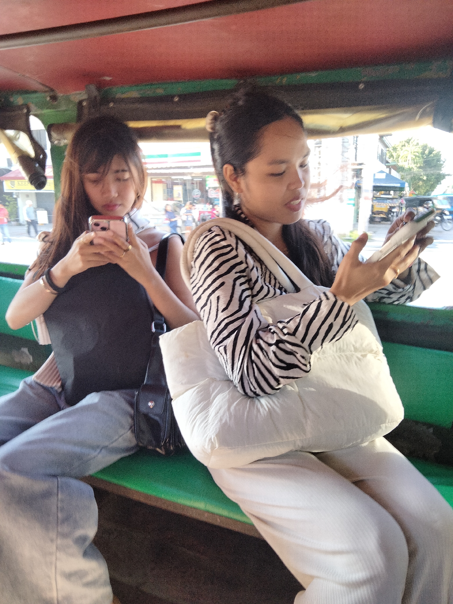 Two young women are seated inside a tricycle in Davao City, Philippines. The woman on the left has long brown hair and is wearing a black tank top, light blue jeans, and a black purse slung over her shoulder. She is looking down at her pink smartphone. The woman on the right is wearing a black and white striped shirt and white pants, with a white shoulder bag. She is also looking at a silver smartphone. The tricycle has a green interior and a red roof. Through the open sides of the tricycle, a street scene is visible with buildings, pedestrians, and other vehicles.