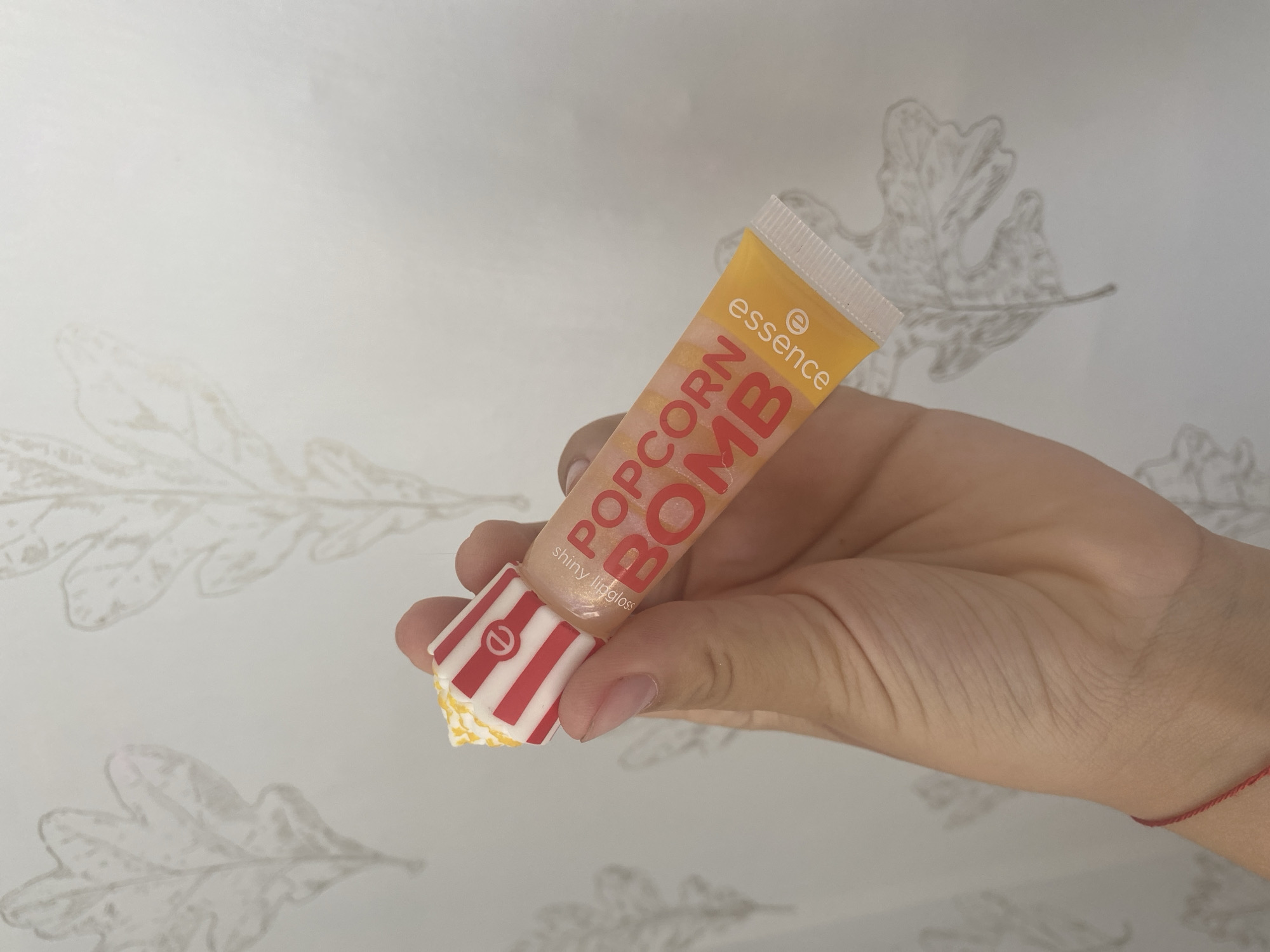 A person's hand holds a lip gloss tube with a popcorn-themed cap against a wall with a repeating leaf pattern. The lip gloss tube is translucent and appears to be peach or orange in color, with "POPCORN BOMB" and "essence" printed on it. The cap is red and white striped, resembling a popcorn box. The hand has a thin red string bracelet on the wrist.