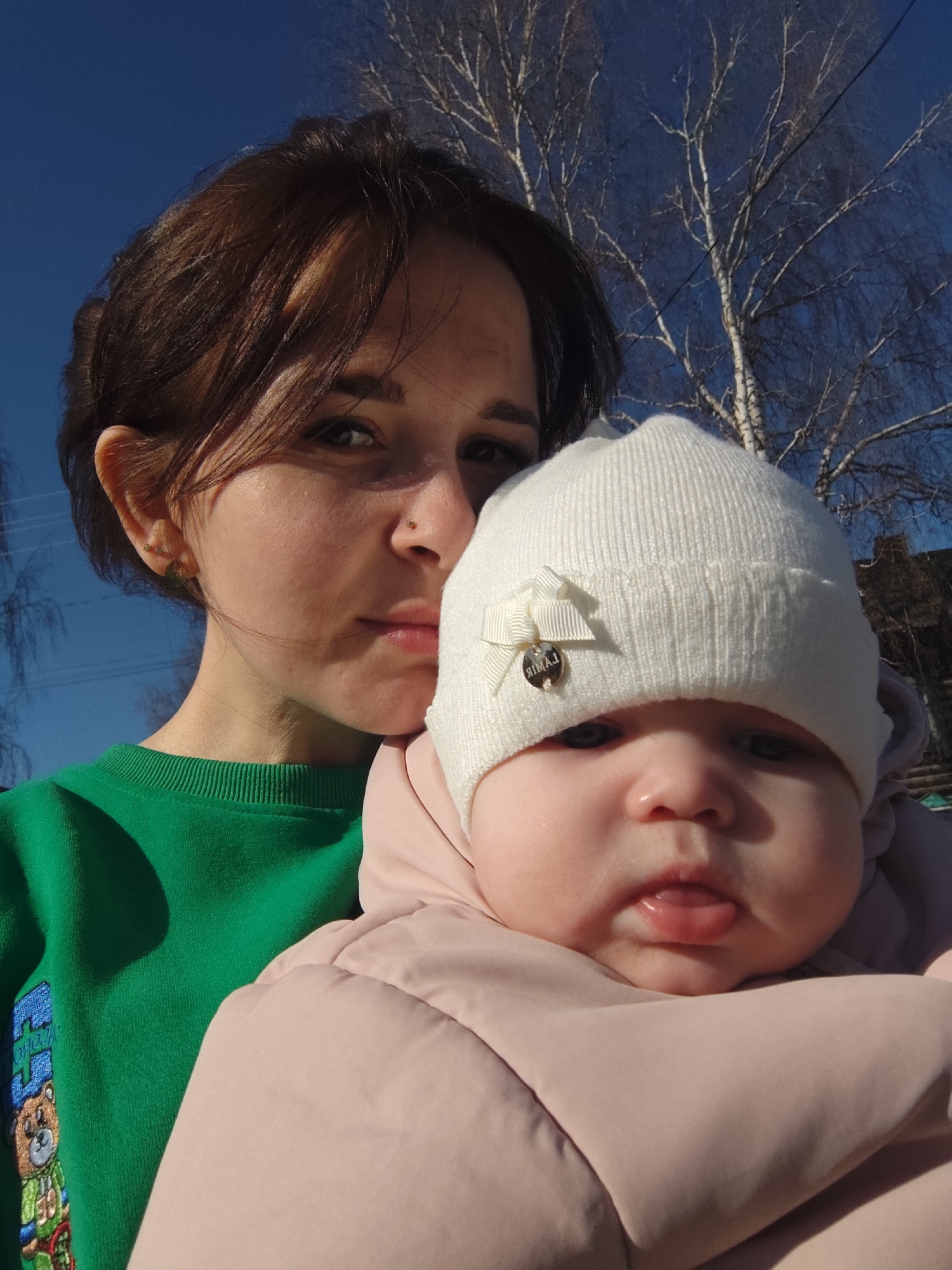 An outdoor, eye-level selfie-style portrait captures a woman holding a baby against a backdrop of a clear blue sky and bare trees, likely birch due to their distinctive light bark. The scene takes place in Фоки, Russia, under bright daylight, suggested by the strong sunlight and clear sky, though the bare trees and warm clothing imply a cooler season like late autumn, winter, or early spring.
The woman, positioned on the left, has dark brown hair, dark eyes, and a small nose piercing. She wears a vibrant green long-sleeved top featuring an embroidered design on the lower left, which appears to include a cartoon bear character. Her expression is neutral to slightly contemplative.
On the right, the baby is held close to the woman. The baby wears a white knit beanie adorned with a small white bow and a metallic charm. The text on the charm reads "ЯМАЯ" (YAMAYA), though it appears mirrored. The baby is dressed in a light pink, possibly padded or puffer, jacket or suit, suggesting protection from the cold. The baby's mouth is slightly open, with its tongue visible, and it looks directly at the camera with a curious or dazed expression.
The bright sunlight casts soft shadows and highlights on their faces and clothing. The overall impression is a tender moment between a mother and her child outdoors.