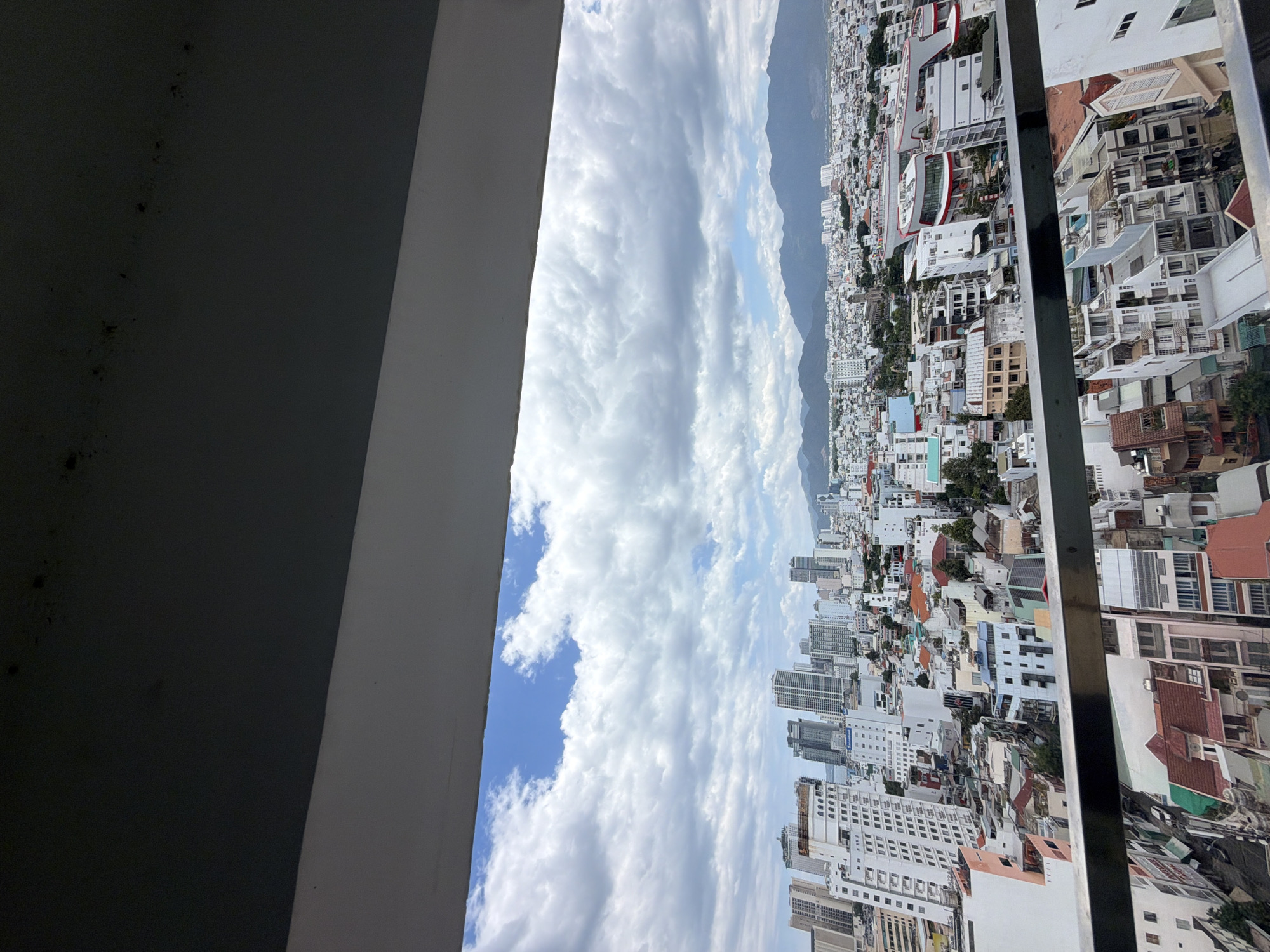This is a high-angle, full shot of a cityscape in Nha Trang, Vietnam, as viewed from a balcony. The foreground is dominated by a dark grey ceiling and a white beam of the balcony structure, obscuring the bottom portion of the view. Below this, the sky is filled with a mix of white and grey clouds against a bright blue background. The mid-ground reveals a dense urban landscape composed of numerous buildings of varying heights and architectural styles, predominantly white and light-colored. Tall, modern skyscrapers stand alongside smaller, older structures with red-tiled roofs. Streets and alleys are visible, densely packed with buildings. In the far background, a mountain range forms a distant horizon, partially obscured by haze. The scene is captured on a cloudy day, with the sunlight filtering through the clouds. There are no people or animals visible in the image.