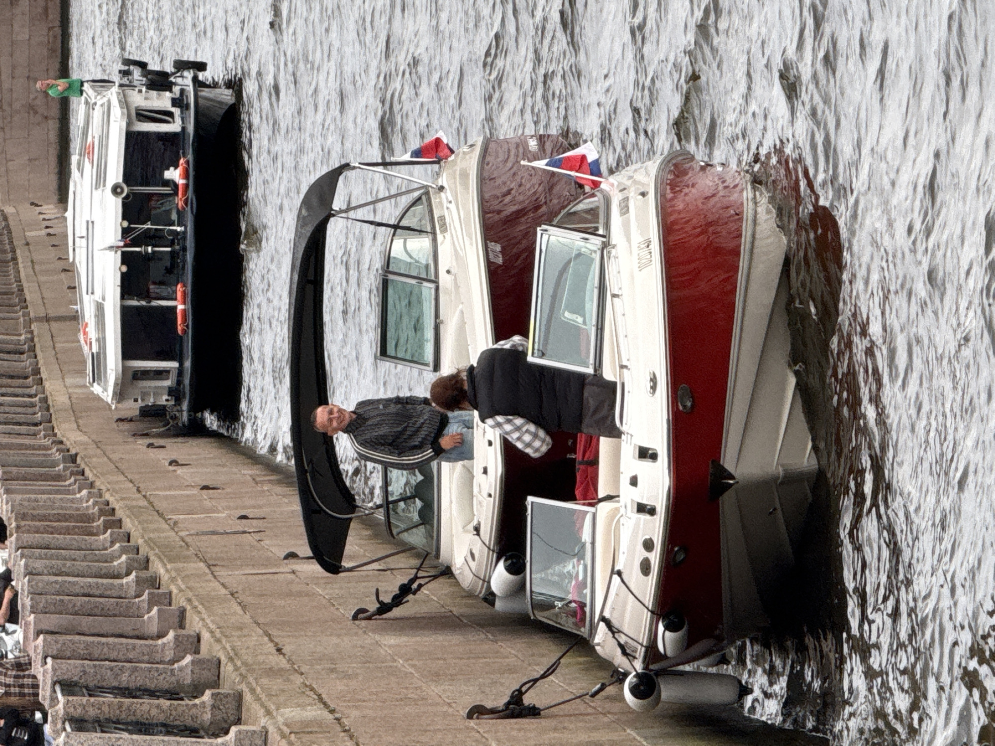 This image shows two boats docked next to a stone embankment with a wide set of stairs leading up from the water. The boats are white with red hulls and have flags flying, suggesting they are in a Russian context, specifically Saint Petersburg.
The first boat, closer to the embankment, is a pleasure craft with a covered cabin. A man in a striped shirt and jeans is leaning over the side, looking towards the camera with a slight smile. Another person, wearing a black vest over a plaid shirt, is also visible inside the cabin.
The second boat, to the right, is partially obscured by the first. It also has a red hull and appears to be a similar style of boat. Both boats are secured with ropes and fenders.
The water is choppy and appears to be a river or canal, typical of Saint Petersburg. The lighting suggests it is daytime, possibly overcast given the lack of strong shadows. The stone embankment and the style of the boats are consistent with a European urban waterfront. There is a person standing on the embankment to the left, wearing a green top. The overall scene depicts a calm day at a dock with people relaxing on or near their boats.