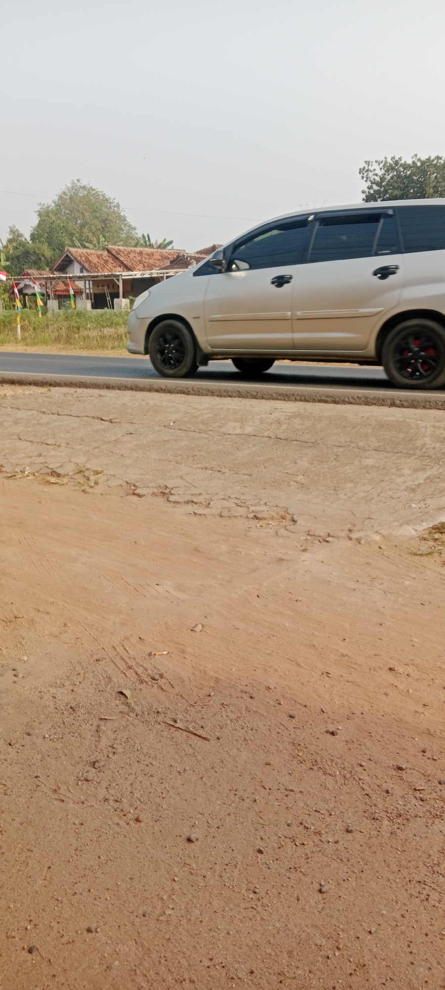 A silver minivan is parked on a paved road in the city of Sidodadi, Indonesia. The minivan has black rims on its wheels. The road is flanked by a dirt shoulder and a grassy area. In the background, there are houses with red tile roofs and trees. The sky is clear and bright.