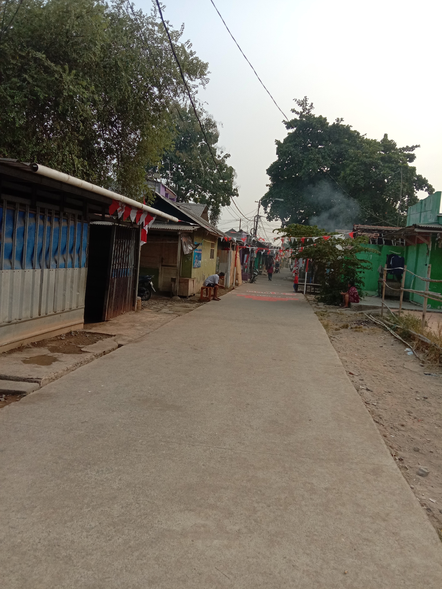 The image depicts a street scene in a densely populated residential area, likely a kampung or informal settlement, within the city of Karawang, Indonesia. The street itself is a paved asphalt or concrete surface, relatively narrow and flanked by simple, functional structures, predominantly houses and small businesses. Many of these buildings appear to be constructed from corrugated metal and wood, with some showing signs of wear.
Along the sides of the street, there are temporary decorations, including strings of small red and white flags, which are the colors of the Indonesian flag. These flags are hung between buildings and over the street, suggesting a festive or commemorative occasion. In some areas, there are also red painted markings on the ground.
There are several people visible in the image. On the left side, a person is sitting on a low stool outside a building with blue and grey corrugated metal siding. Further down the street, on the right side, another person is seated on the ground, leaning against a tree or bush, wearing a red and white patterned garment. In the middle distance, several individuals are walking along the street, and a child is standing near the center. A motorcycle is parked on the left side of the street.
The environment is characterized by the presence of mature trees, particularly on the left and right edges of the frame. There is a noticeable haze or smog in the background, obscuring the sky and distant structures, which could be indicative of air pollution or dust. Smoke is also visible rising from behind a cluster of trees in the middle ground. The ground next to the paved street on the right appears to be unpaved, consisting of dirt and gravel, with some scattered debris. The overall impression is one of a vibrant, lived-in community, possibly celebrating a national holiday or event.