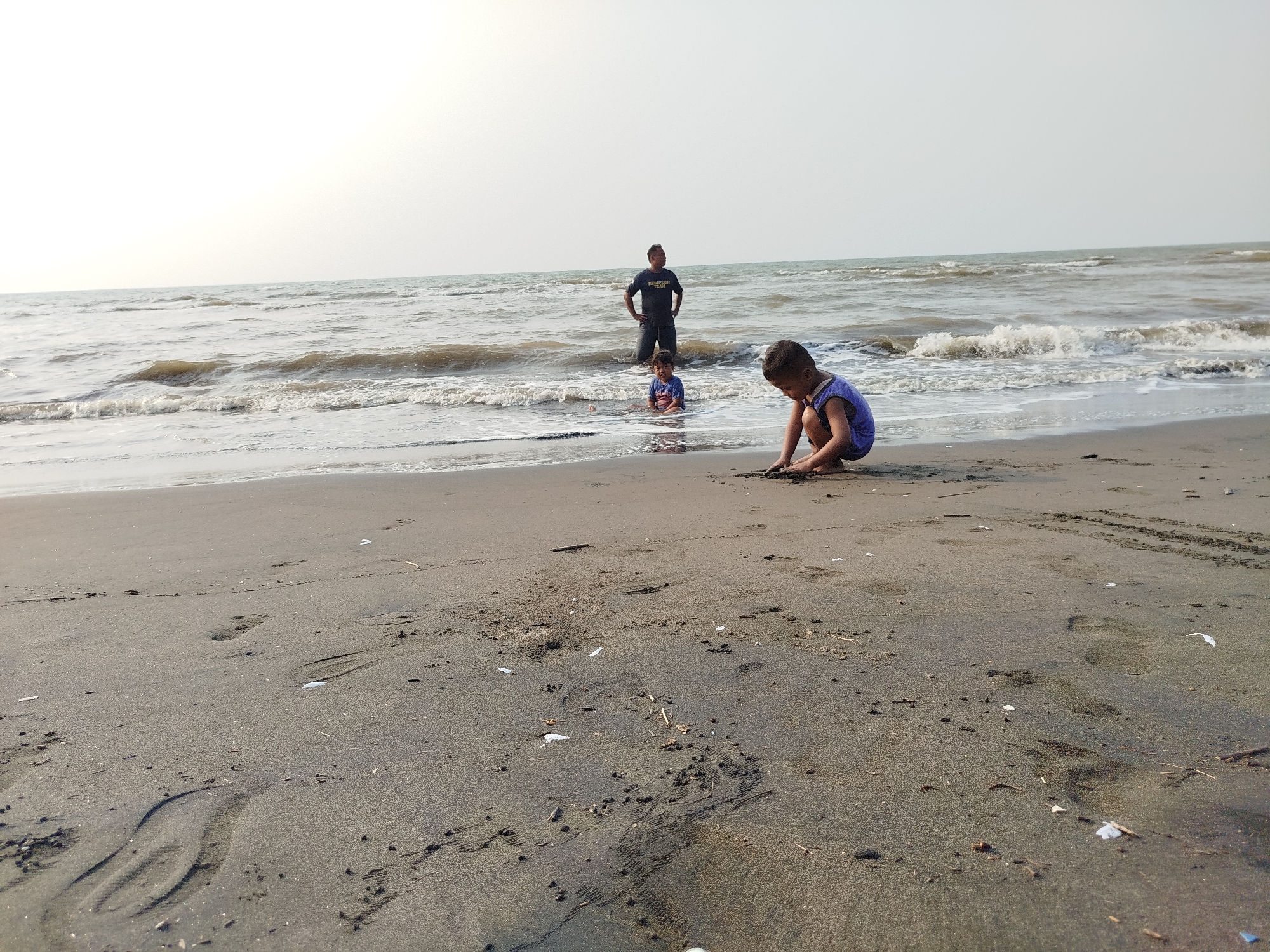 A medium shot captures a family enjoying a beach in Jungsemi, Indonesia. The foreground is dominated by dark, wet sand with footprints and small debris. In the midground, a young boy in a blue sleeveless shirt squats on the sand, actively playing with it. Further back, a second child sits in the shallow water, looking towards the man. Behind them, a man stands in the ocean, facing away from the camera with his hands on his hips. The sea is calm with gentle waves, and the sky is a pale, hazy white. The overall scene depicts a serene family outing at the beach.
