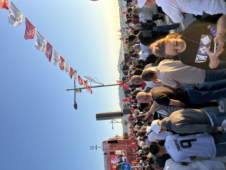 The scene is a crowded public square in İzmir, Türkiye (latitude: 38.437, longitude: 27.141), likely Konak Square. A large crowd of people are gathered, many of whom are holding red and white flags. The flags are flying in the breeze, while a speaker is addressing the crowd from a stage in the background. The people are listening intently, some holding phones and others making small talk with people nearby. There are some trees and a large building behind the crowd. The sky is blue with a few fluffy white clouds. It is a bright, sunny day.