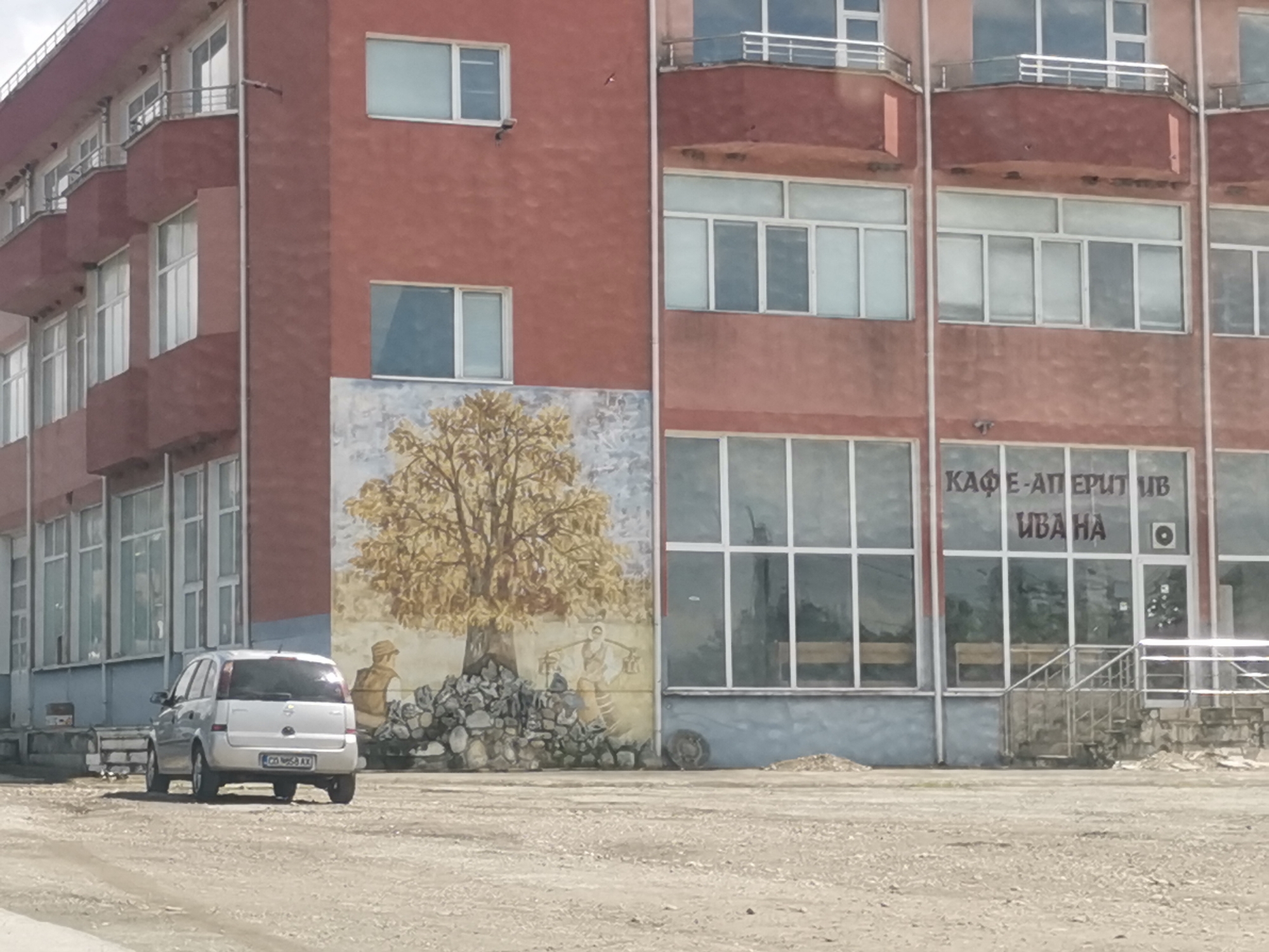 The image captures a building in Mirovo, Bulgaria, featuring a large mural and a parked car. The building consists of two main sections. The left section, painted in a reddish-brown, has several floors with balconies and numerous white-framed windows. A large mural dominates the lower part of this section, depicting a golden-leafed tree with two figures: one seated near a pile of stones, and another carrying a yoke with baskets, evoking an agricultural or pastoral theme.
The right section of the building, with a slightly lighter reddish facade, features extensive ground-floor windows, indicating a commercial establishment. Through these windows, Cyrillic text is visible, reading "КАФЕ-АПЕРИТИВ ИВАНА," which translates to "CAFE-APERITIV IVANA." An entrance with steps and a metal railing leads into this commercial space.
In the foreground, a silver compact car, identified by its Bulgarian license plate "CO 9568 AX" (indicating Sofia registration), is parked in front of the mural, facing away from the viewer. The ground in front of the building is unpaved, consisting of dirt and gravel. The scene is set during the daytime, under bright, possibly overcast conditions, with no specific activity taking place other than the car being parked. The architecture and signage provide contextual cues for a Bulgarian setting.