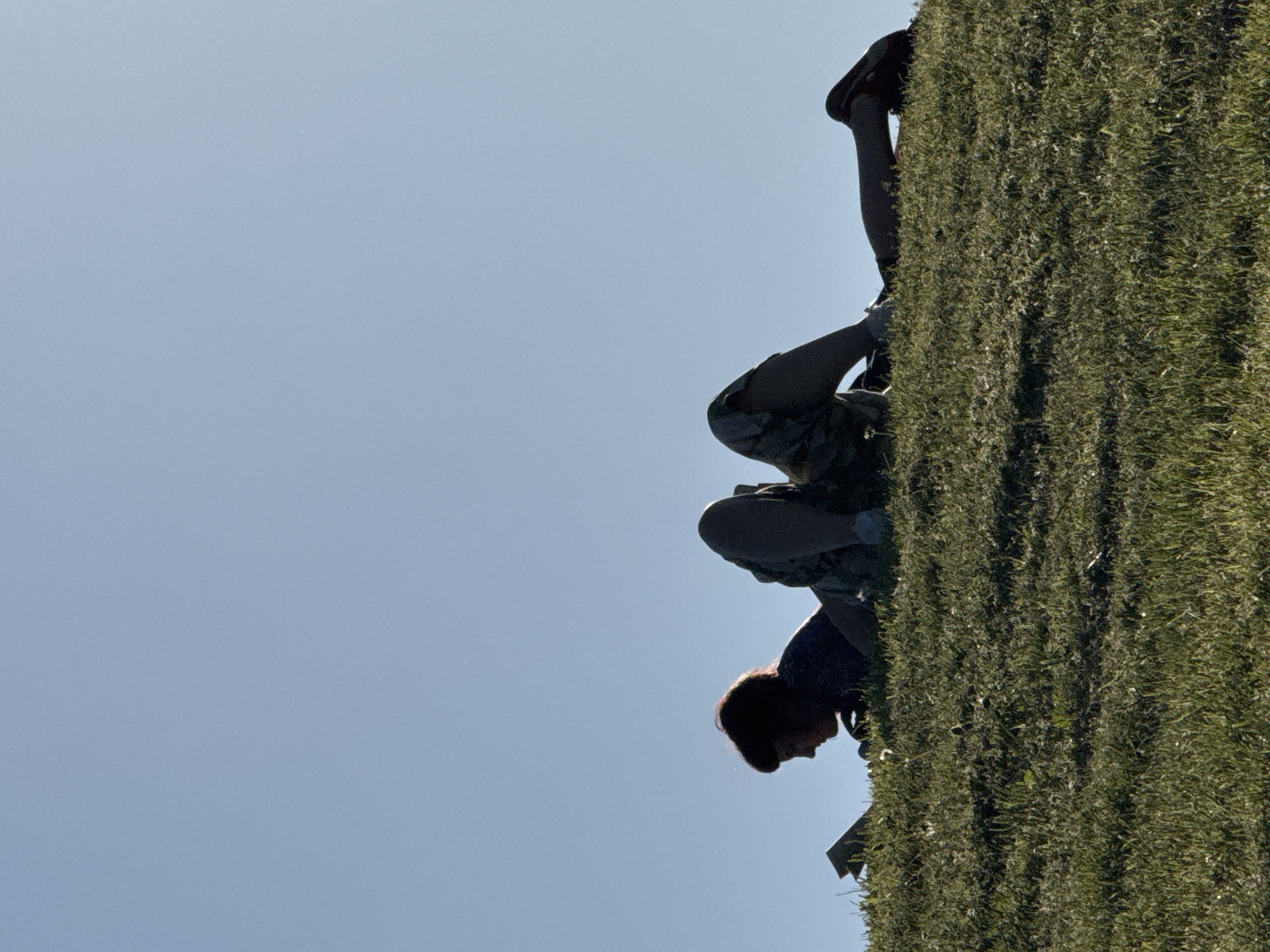 A person climbs a grassy wall in Sofia, Bulgaria. The wall is densely covered in green vegetation. The person is wearing dark shoes, patterned shorts, and a dark shirt. The sky is pale blue and mostly clear.