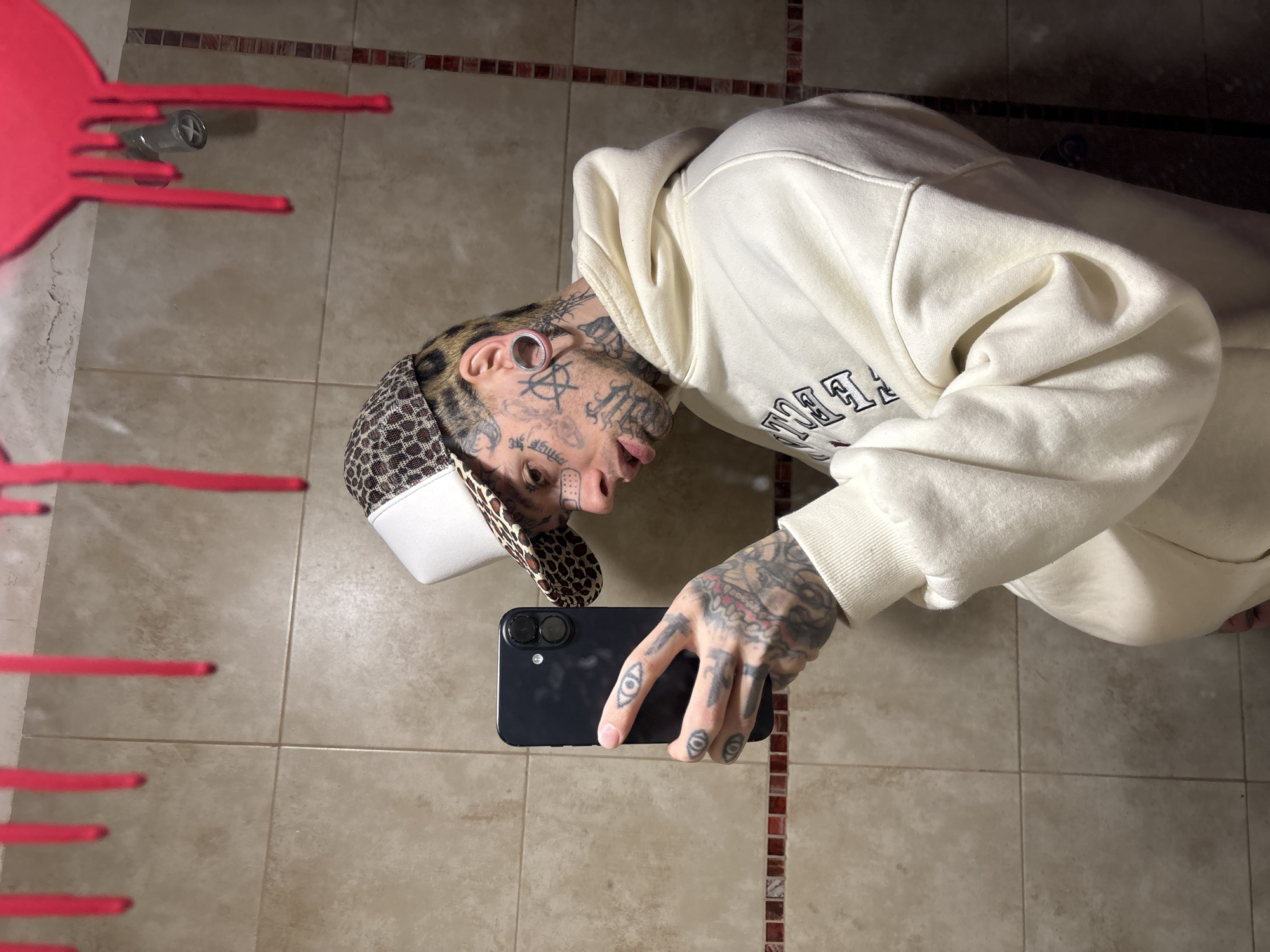 A man is taking a selfie in what appears to be a bathroom. He has numerous tattoos on his face and hands, and is wearing a white hooded sweatshirt and a baseball cap with a white front and leopard-print brim. He is holding a black smartphone. To the left of the man is a red object with silver faucet, which resembles an artistic sculpture. The bathroom has tan tiles on the floor and wall. The location is not evident from the image.