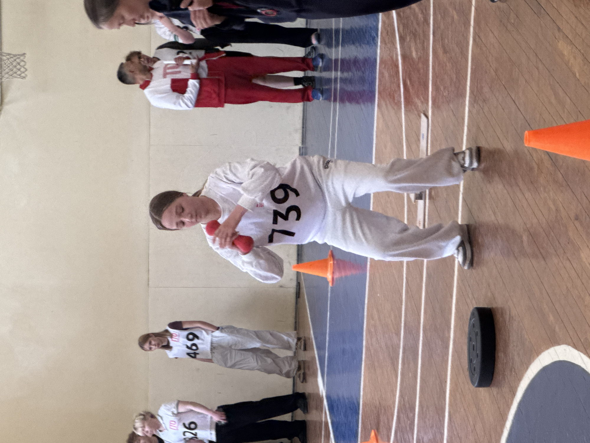 The image shows an indoor athletic event. A young woman wearing a white tracksuit with the number 739 is in the foreground, holding a red baton. She is in motion, participating in a relay race or similar competition. Other participants wearing numbered bibs (e.g., 469, 826) are visible in the background. Orange cones mark the lanes on a wooden gymnasium floor. A black hockey puck is also on the floor. A blue and white court marking is visible, possibly for basketball. A basketball hoop is attached to the wall. The scene is likely an indoor sports arena or school gymnasium. The exact location in Perm, Russia cannot be determined from the image.
