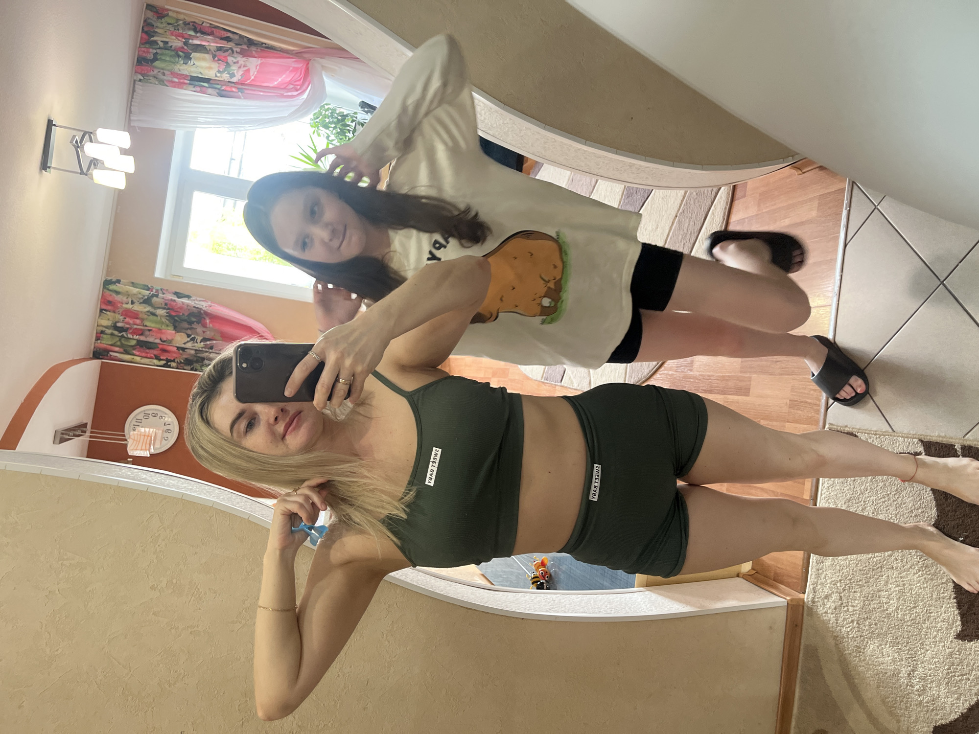 Two females are visible in a mirror. One female is holding a phone and taking a photo, while the second female is behind her and to the right. The female holding the phone is wearing an olive green matching sports bra and shorts set. The second female is wearing a light-colored long-sleeved shirt with a cartoon image and black shorts. Flooring varies from tile to carpet to wood laminate. Curtains and windows are visible in the background. The location is Chaikovsky, Russia.