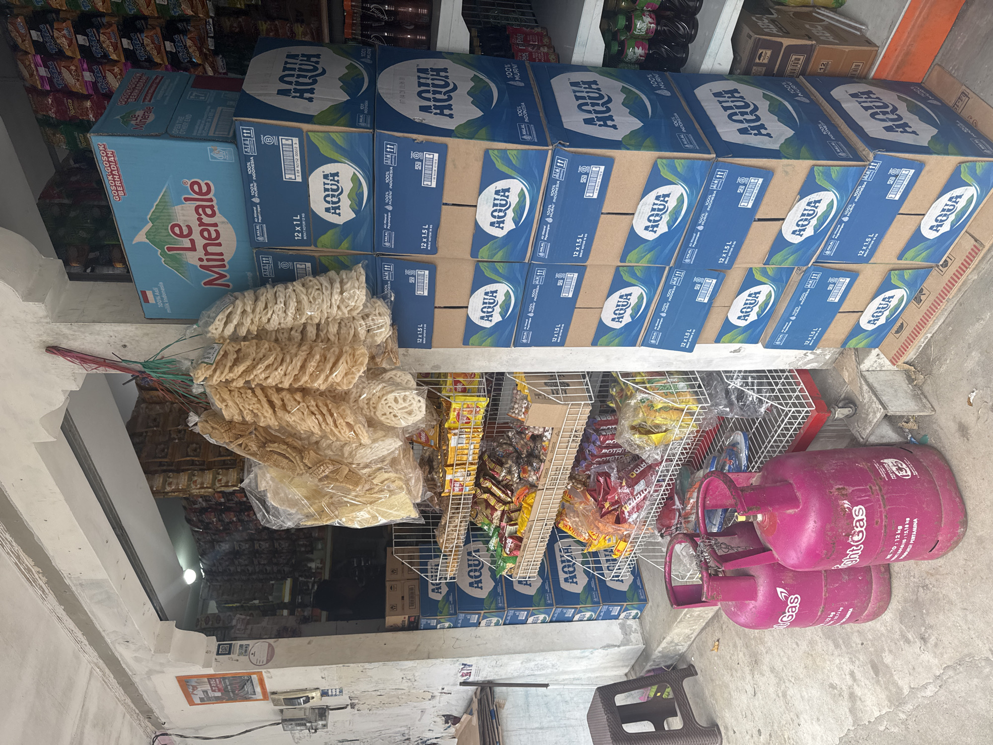 The image depicts the interior of a small shop in North Kuta, Indonesia. The shop is stocked with various items for sale. Cases of "Aqua" bottled water are prominently stacked along the top and lower part of the shelves. Beside the water, there is a blue case of "Le Minerale" mineral water. There are pink gas cylinders for household cooking. A wire rack displays smaller items such as snacks and candies. Bags of Indonesian crackers "Kerupuk" hang from the top. The store appears to be located indoors.